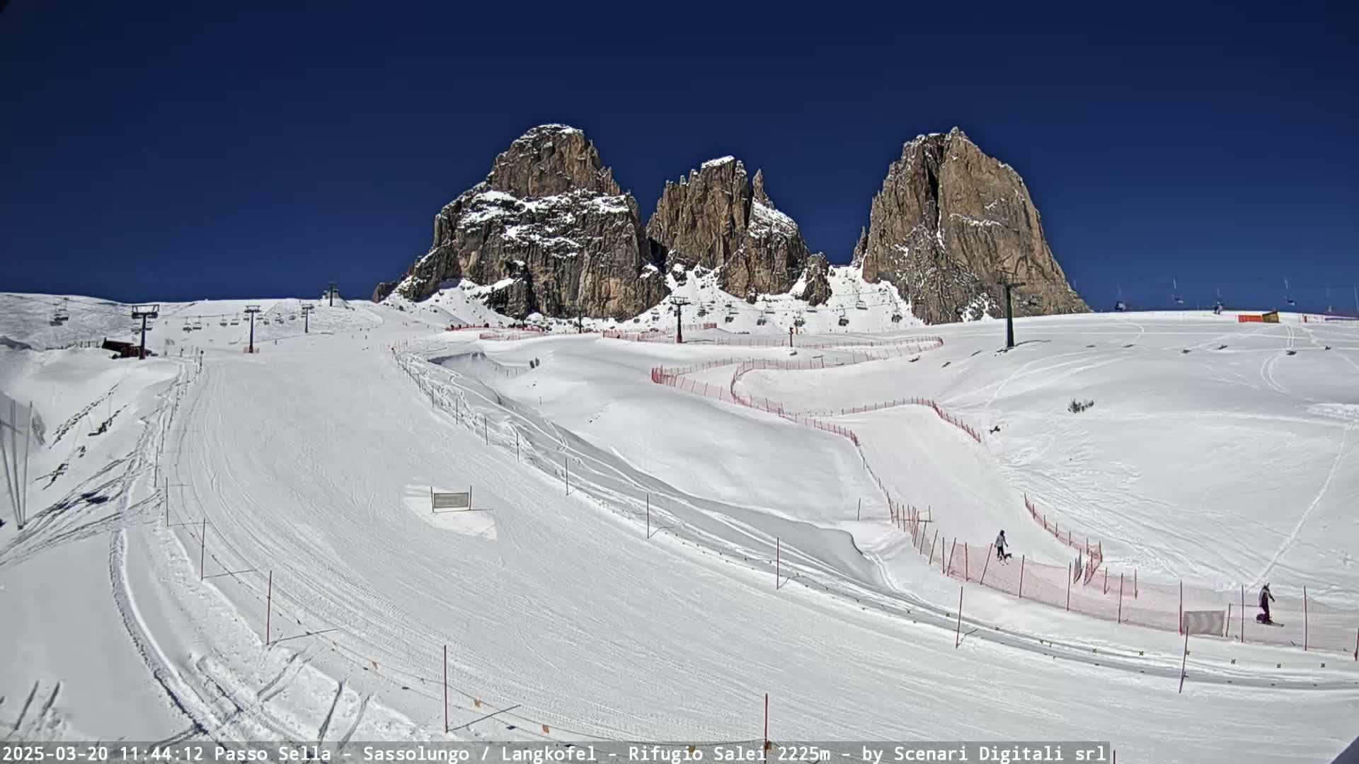 A snow-covered ski slope with several ski lifts and rocky mountain peaks in the background under a clear blue sky.