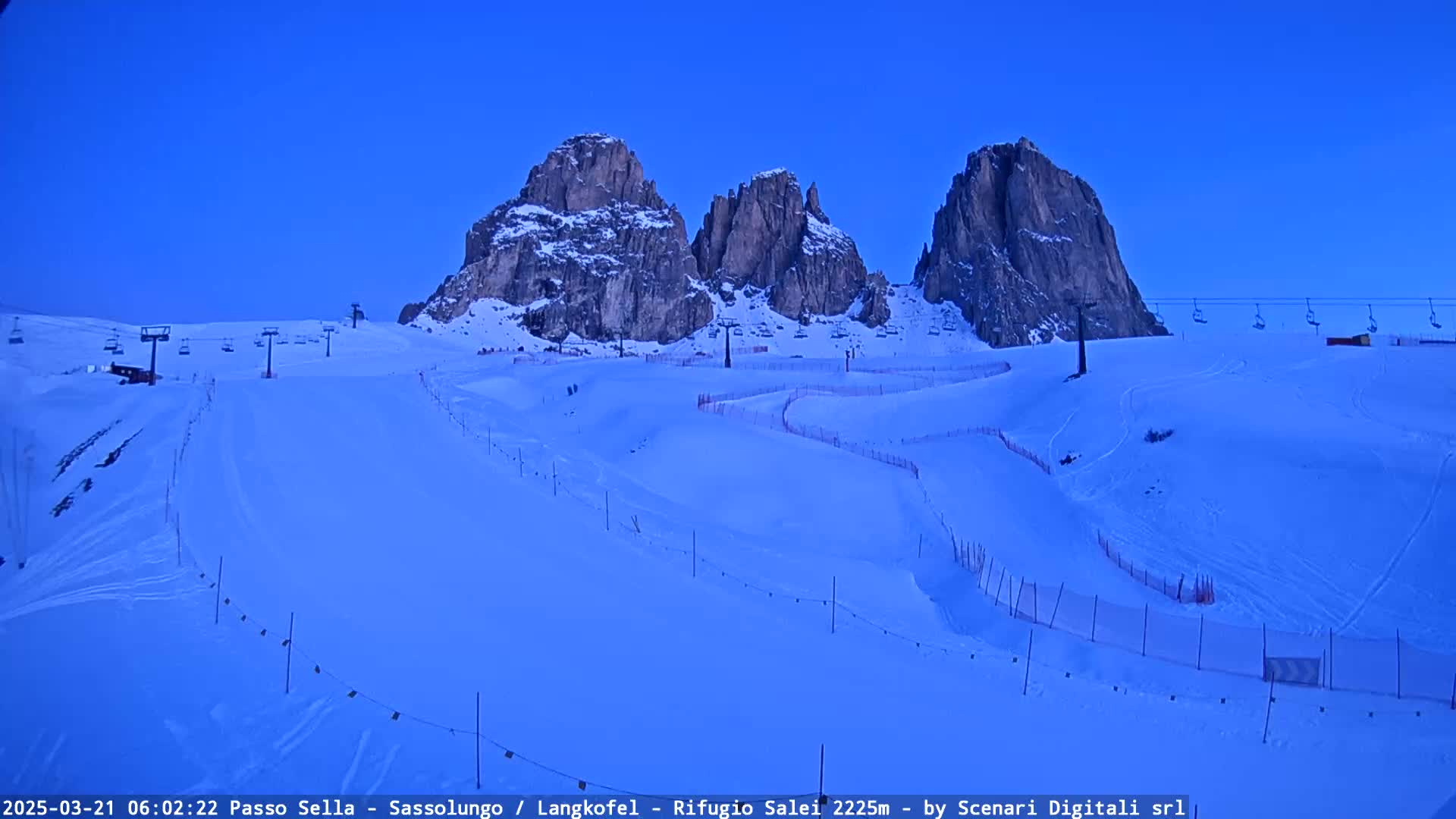 A snow-covered ski slope with chairlifts and tall, rocky mountains in the background under a clear, twilight blue sky.
