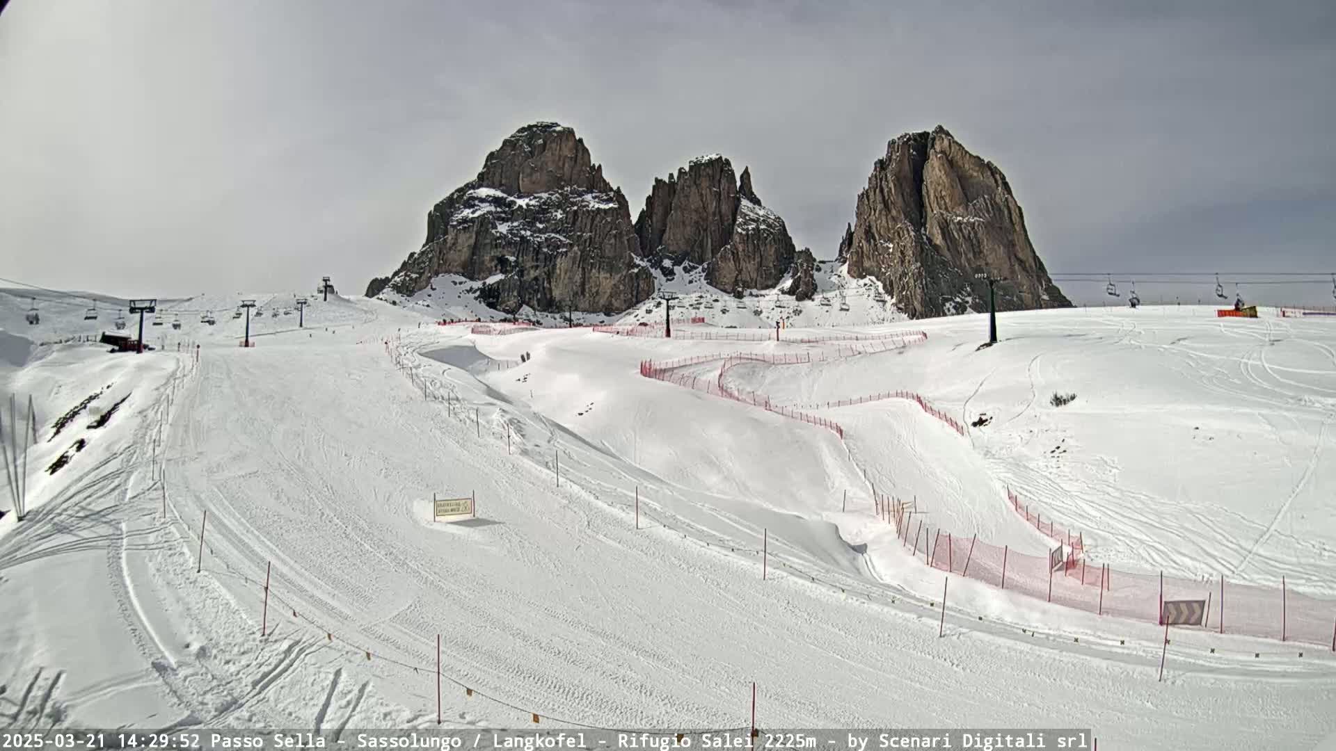 A snow-covered ski slope with several ski lifts and tall rock formations in the background under a cloudy sky.