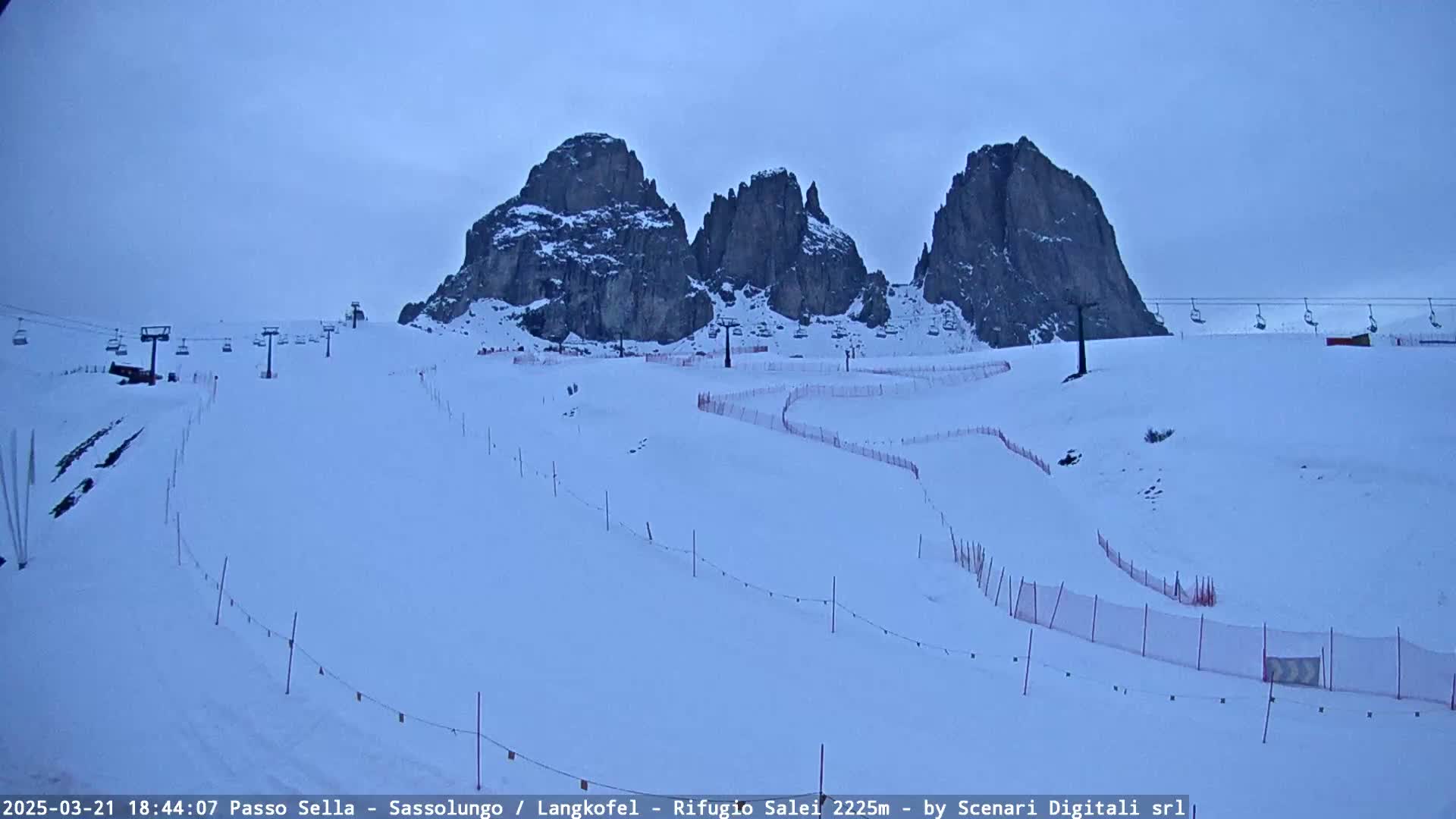 A snow-covered ski slope with chairlifts and fencing winds around several rocky peaks under an overcast sky.