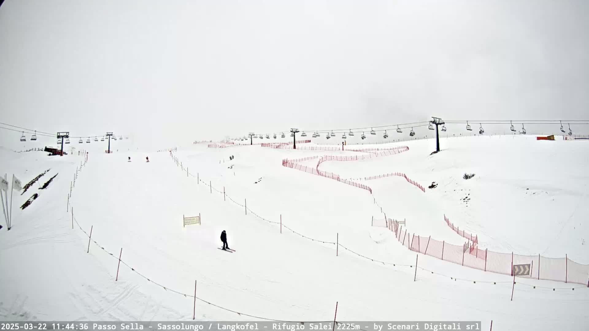 A snowy ski slope with chairlifts and a few skiers visible under overcast skies.