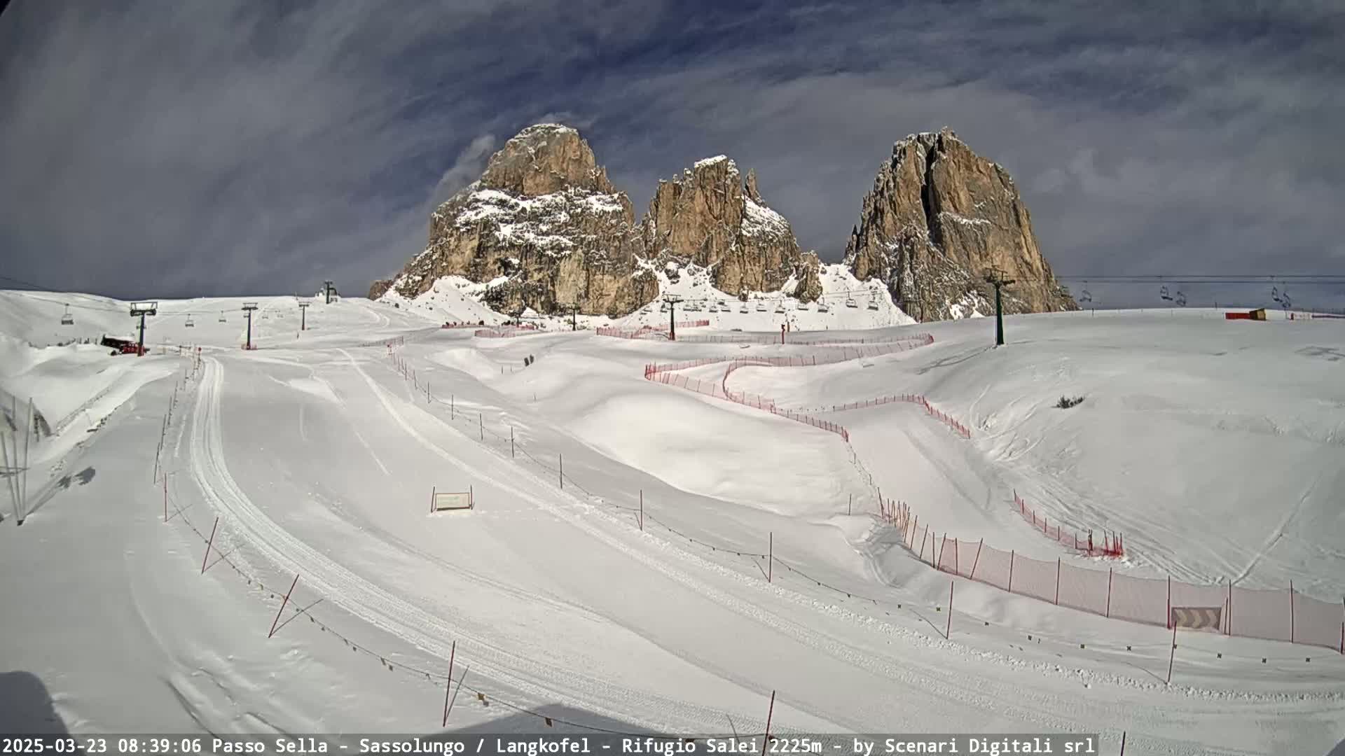 A snow-covered ski slope with several runs, ski lifts, and rock formations in the background under partly cloudy skies.