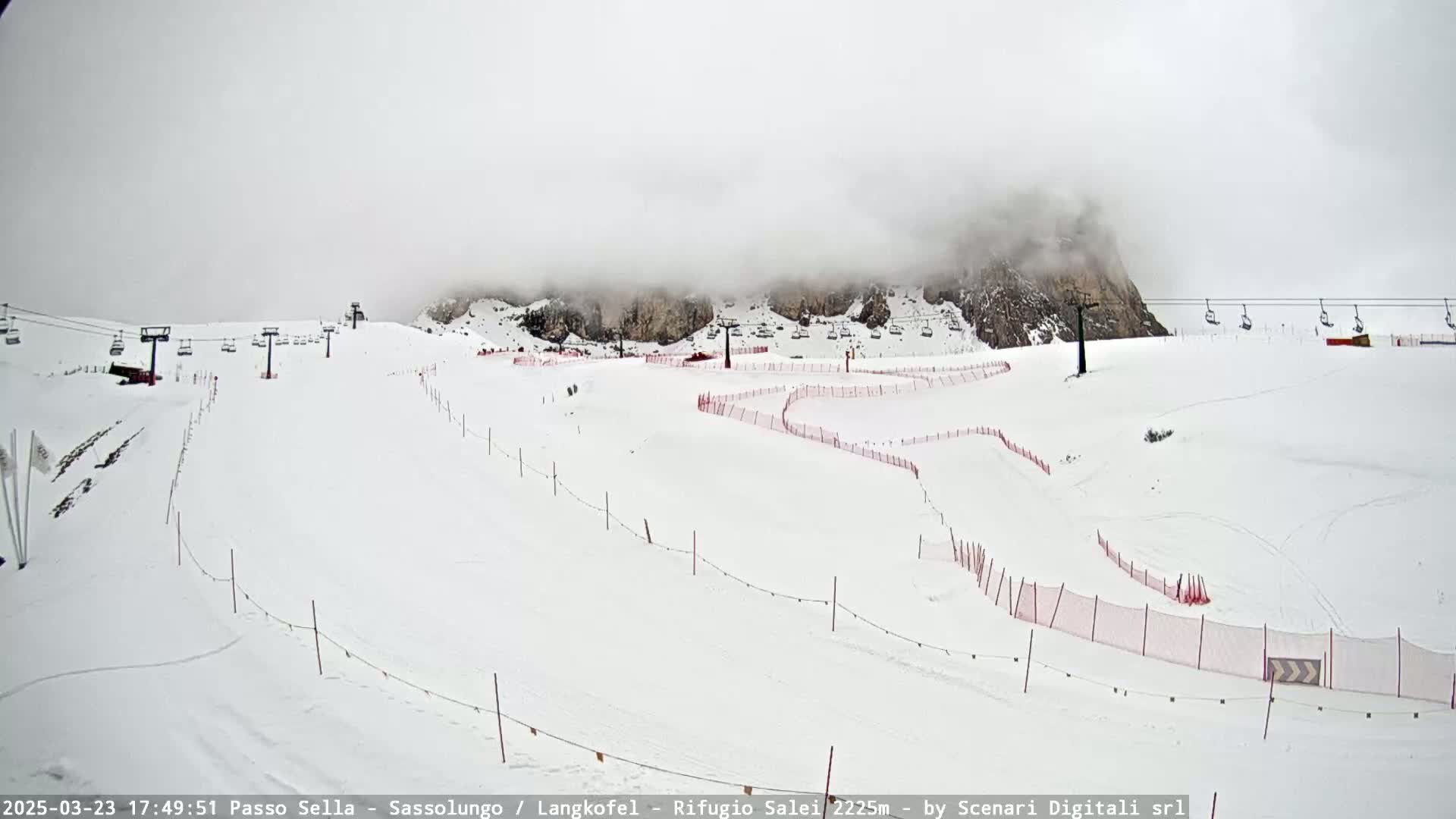 A snowy mountain ski slope with chairlifts and red safety nets is partially obscured by fog.