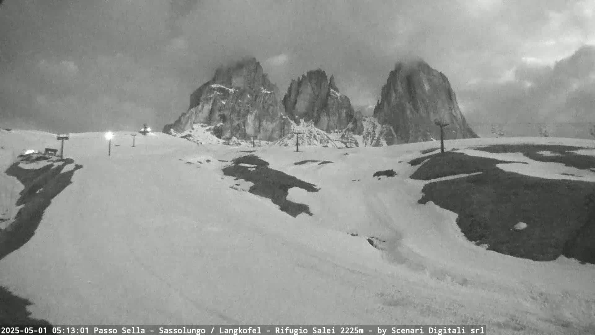 A snow-covered mountain landscape with several rocky peaks under a cloudy, overcast sky, showing some ski lift infrastructure.
