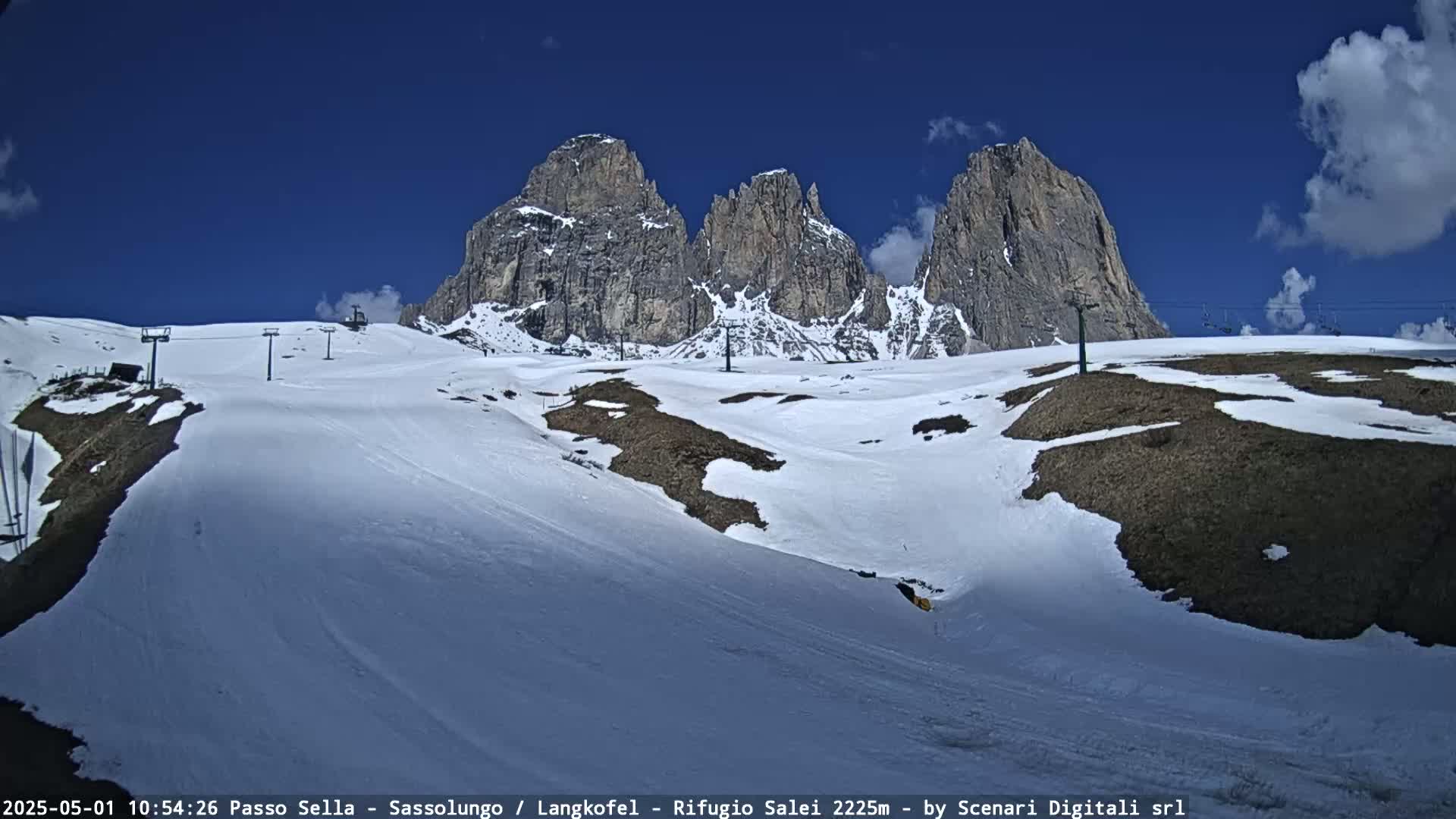 A snow-covered mountain slope with ski lifts and patches of brown earth, under a mostly clear blue sky with a few clouds, features a dramatic backdrop of tall, rocky peaks.
