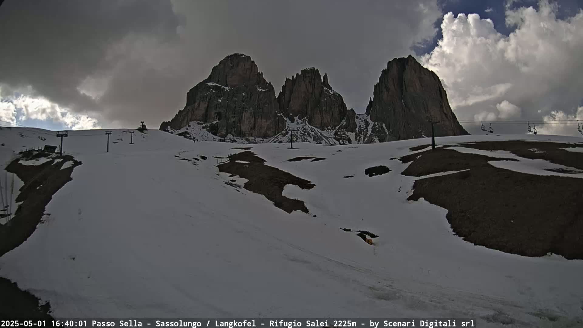 A snow-covered mountain landscape with dark, cloudy skies features a ski lift and several rocky peaks in the background.