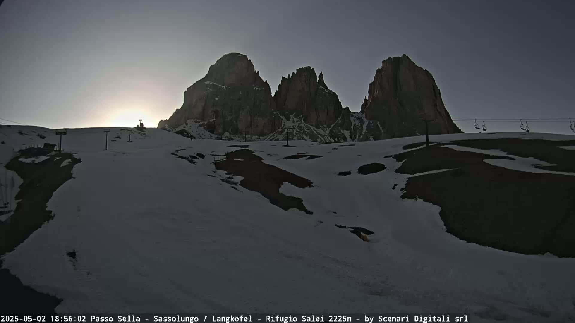 A snowy mountain landscape shows a setting sun behind three prominent rock formations, with empty chairlifts visible in the foreground.