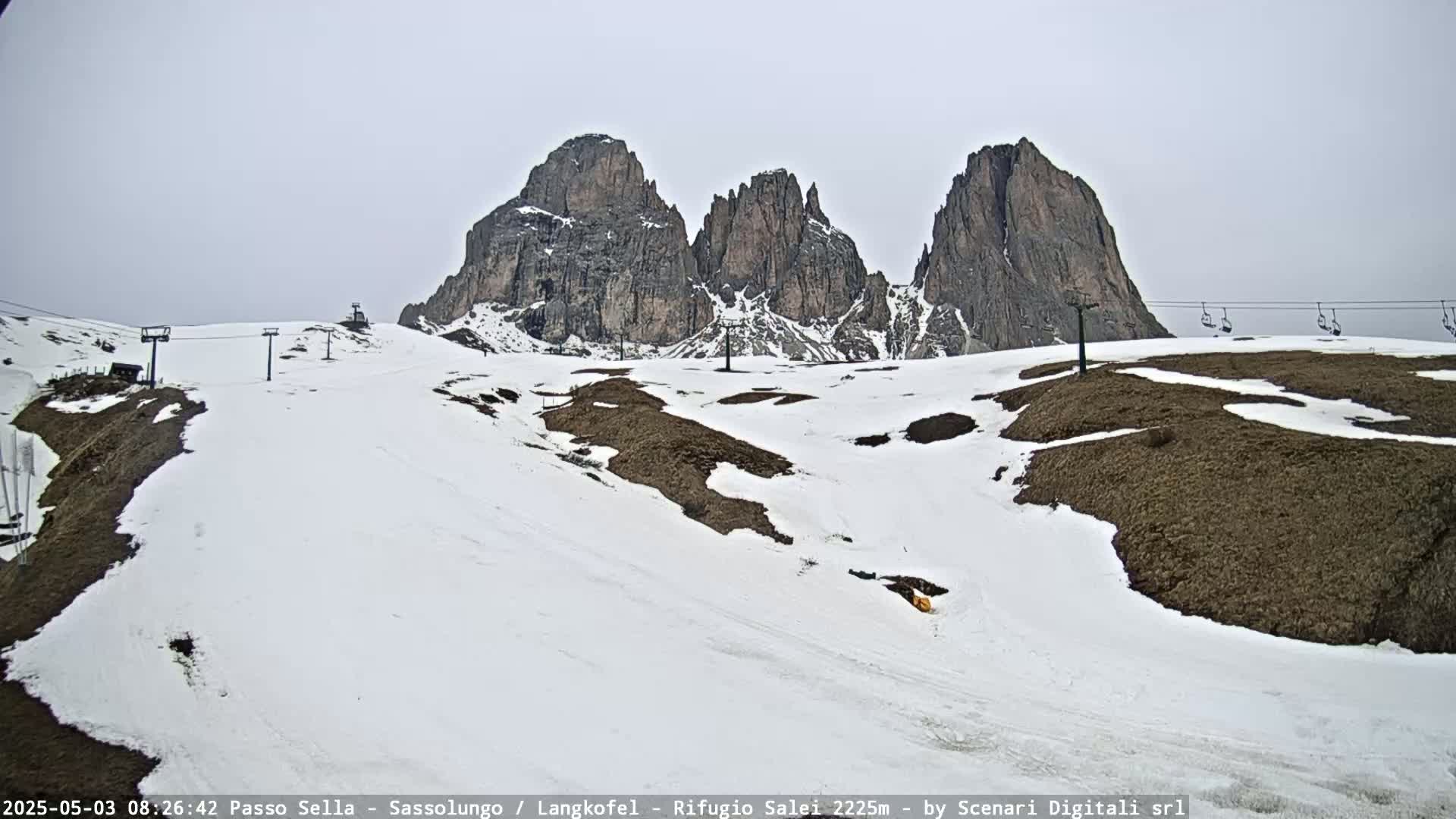A snow-covered mountain slope with ski lifts and three prominent rock formations in the background under an overcast sky.