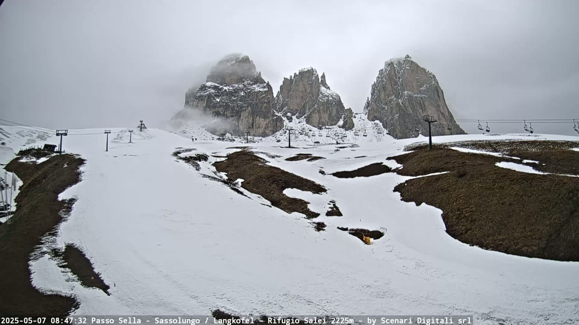A snowy mountain landscape with partially cloudy skies shows several chairlifts and rocky peaks partially obscured by fog.