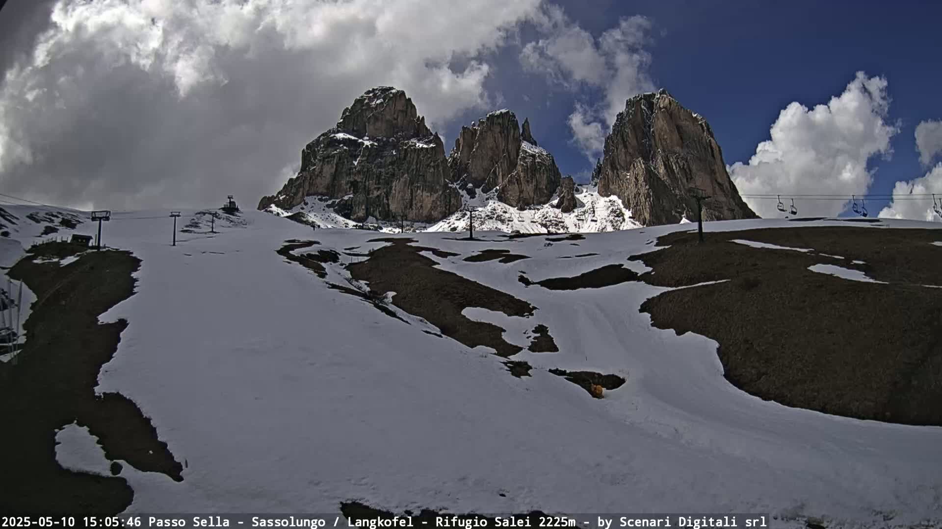 A partly cloudy day shows snow-dusted hills and mountains with chairlifts and a few rocky peaks under a blue sky.