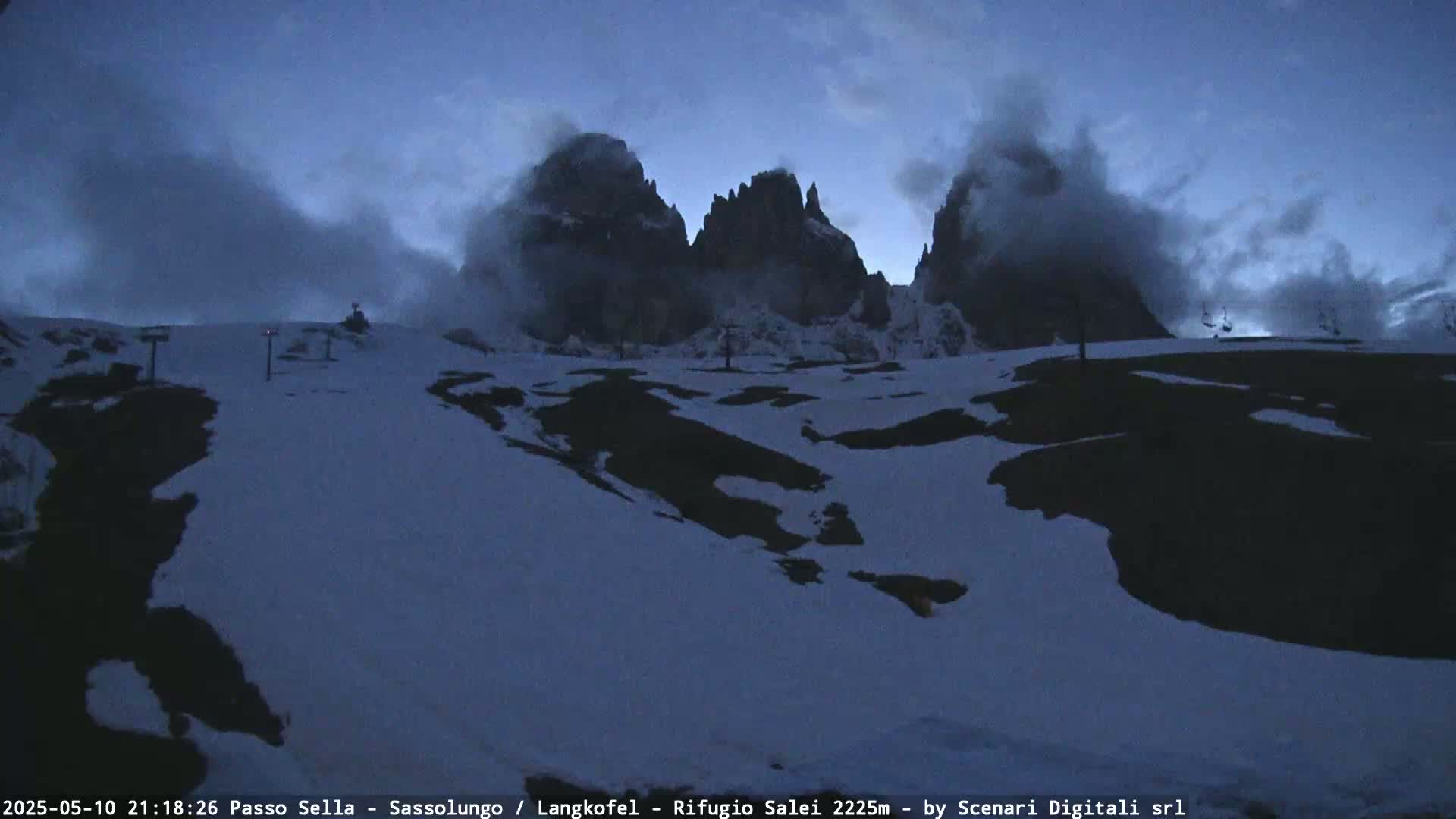 A snow-covered mountain landscape at dusk, with partially cloudy skies and the silhouettes of tall rock formations in the background.