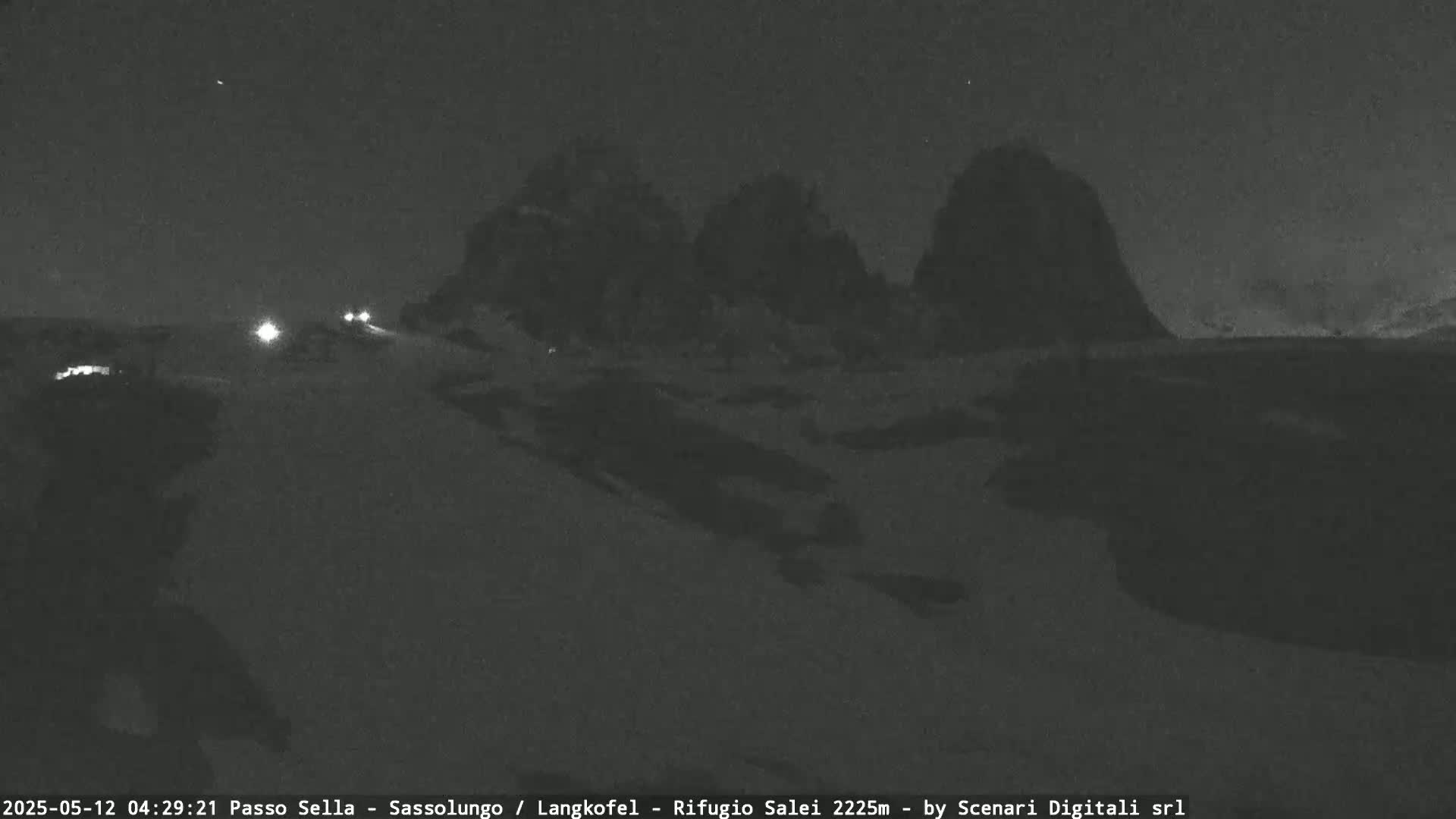 A nighttime view of a snow-covered landscape with dark, mountainous silhouettes in the background, and a few vehicle headlights visible on a road in the foreground.