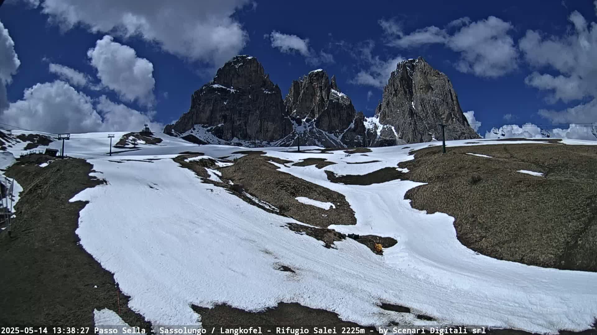 A partly snow-covered mountain landscape under a partly cloudy blue sky shows several jagged peaks in the background.