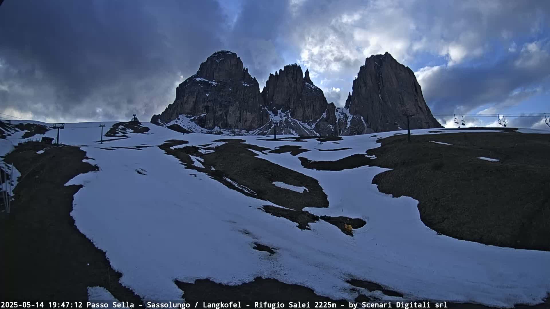 A partially snow-covered mountain landscape with a cloudy sky features a ski lift and tall, dark rock formations in the background.