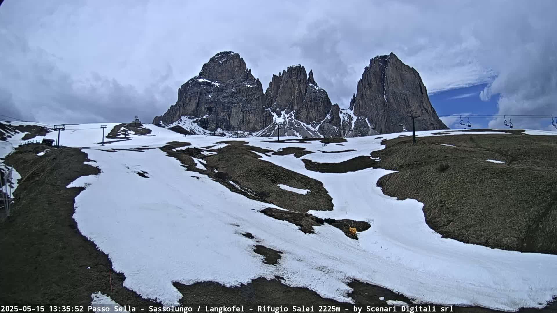 Partially snow-covered ground slopes gently downward toward a viewer, with a backdrop of three towering, rocky peaks under a partly cloudy sky.