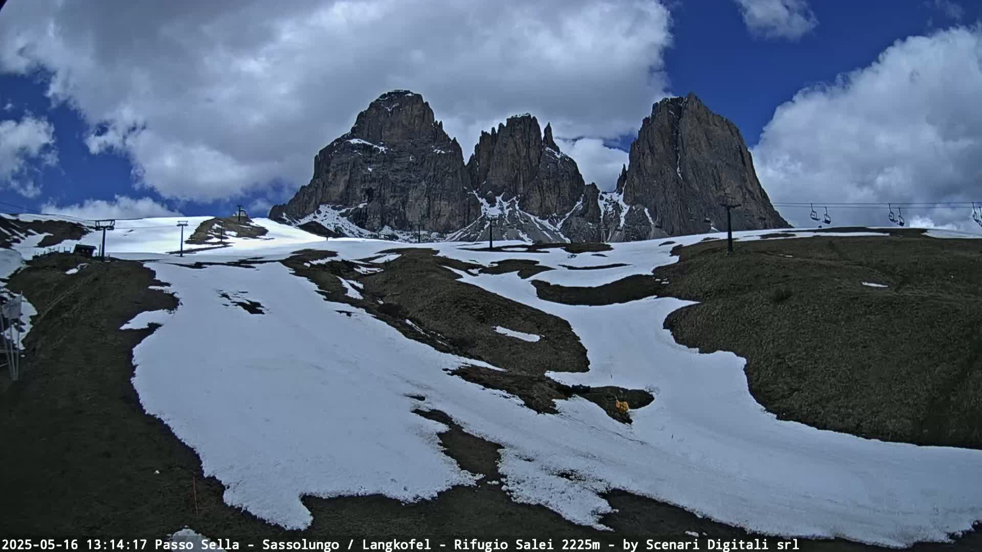 A partly cloudy sky overlooks a snow-dusted mountainside with patches of dark brown earth, featuring a chairlift and a range of jagged, dark peaks in the background.