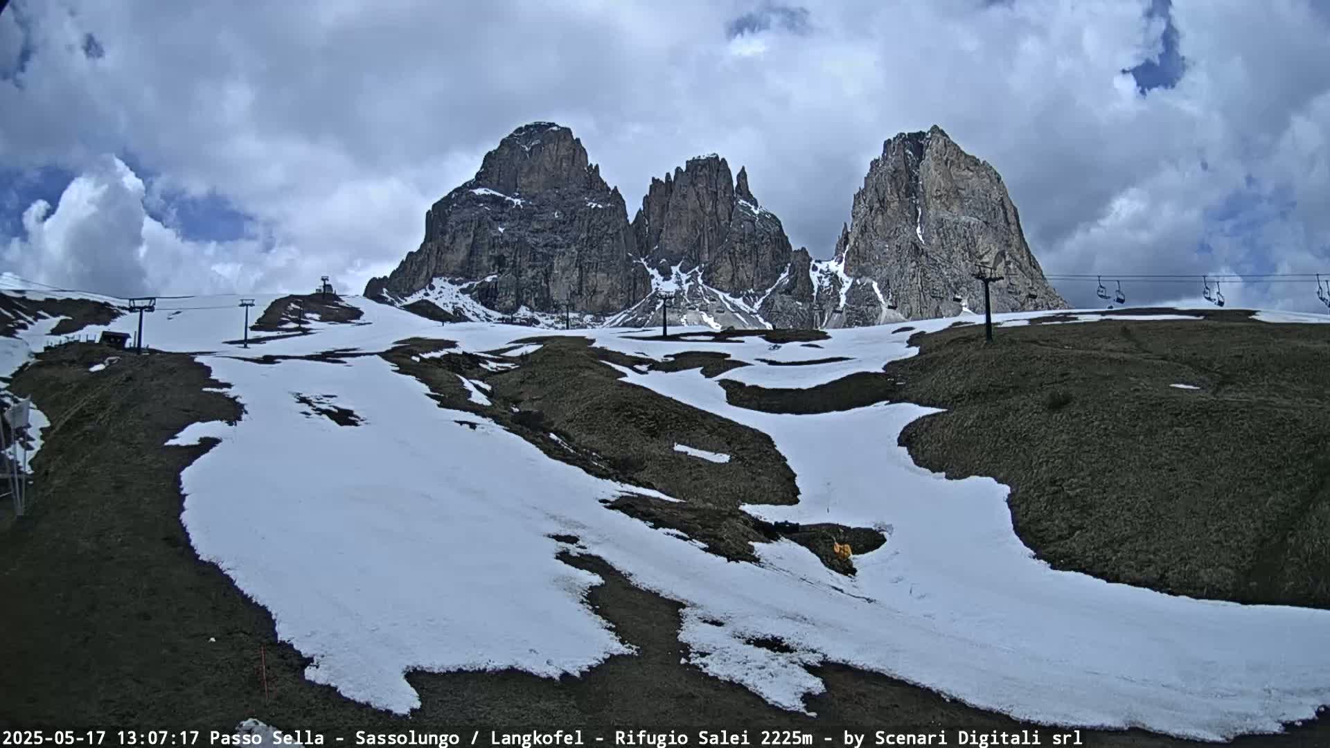 A partly cloudy day reveals a mountainous landscape with patches of snow, ski lifts, and dark-colored, rocky hills.