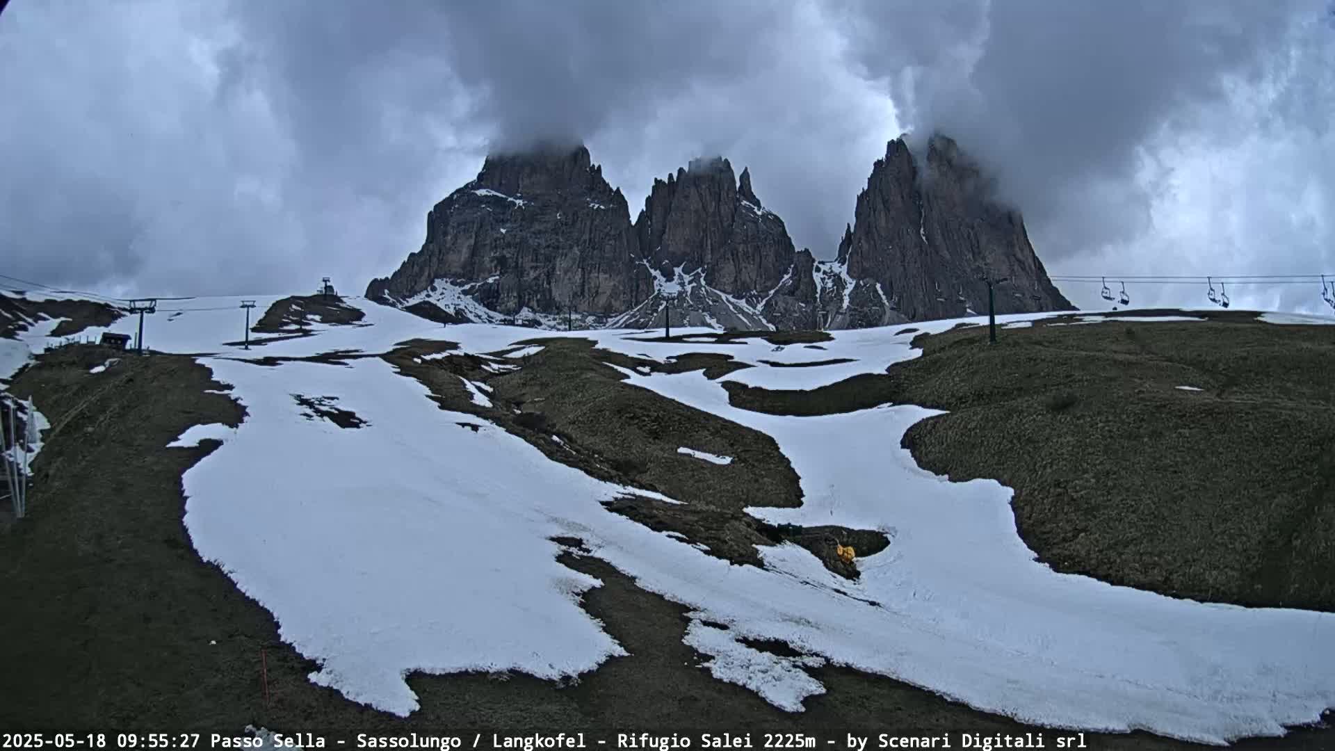 A mountain landscape with patches of snow under a cloudy sky shows a ski lift and several prominent rock formations in the background.