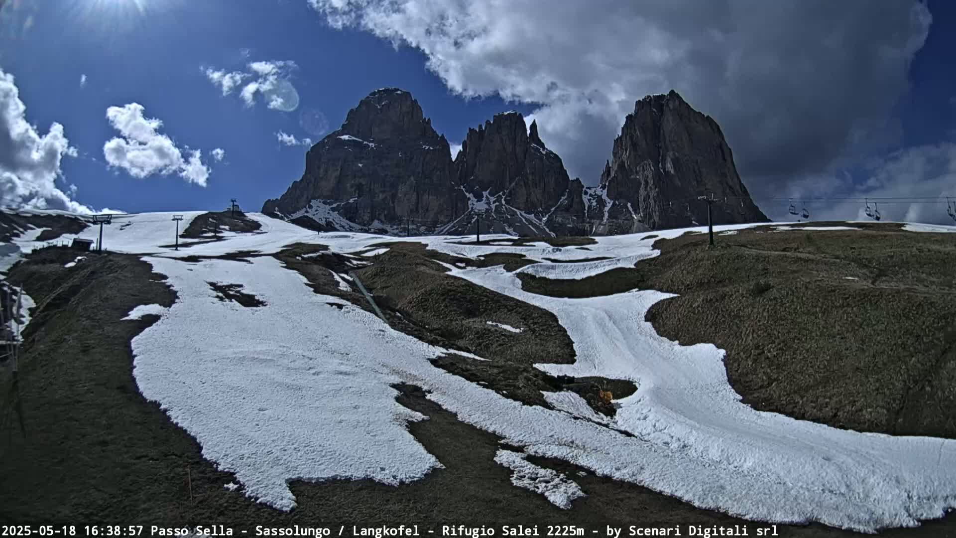 A partly sunny day shows patches of snow on a mountainside with several ski lifts and a background of jagged, dark peaks.