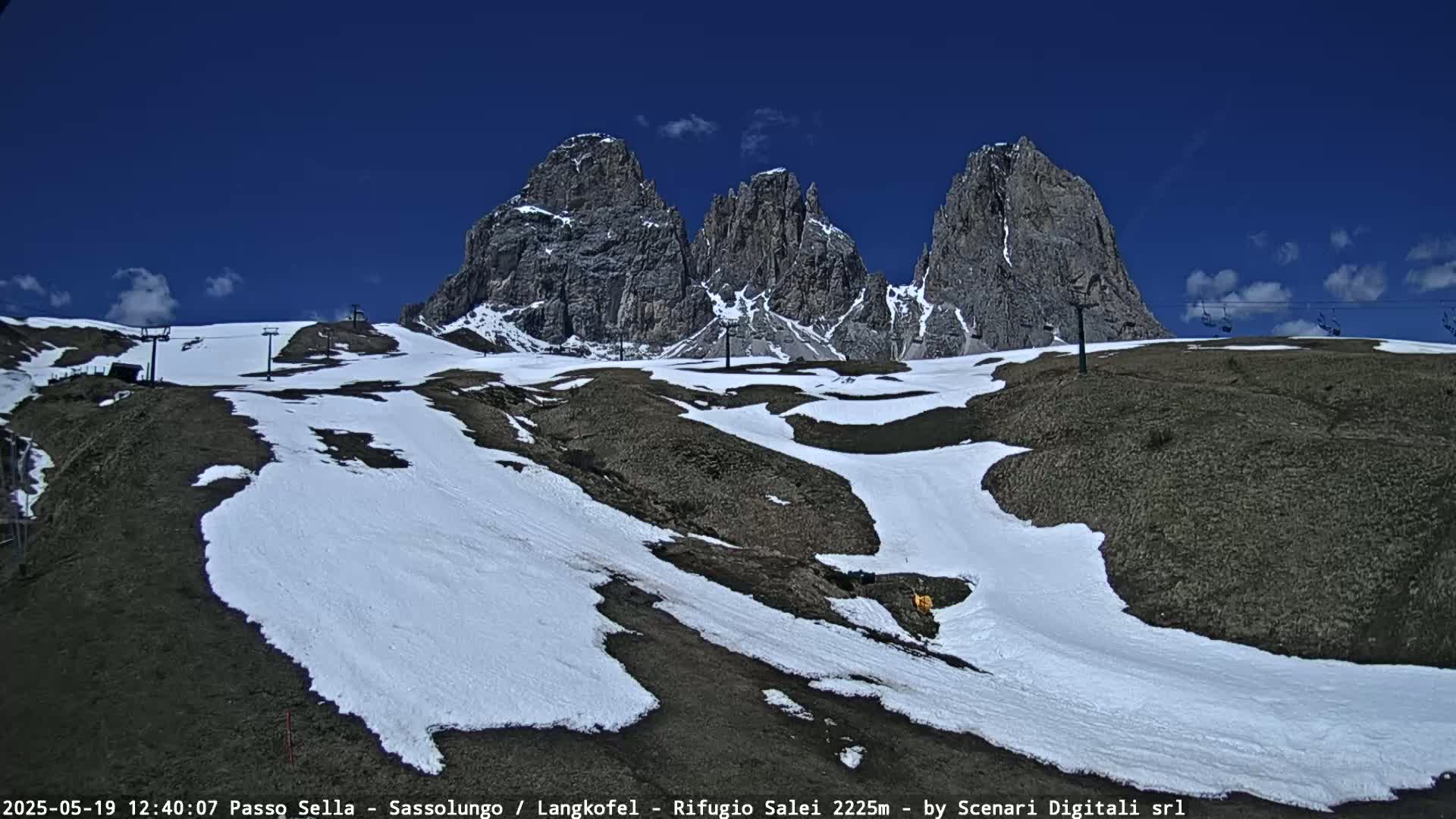 A mountain landscape with patches of snow on the ground under a clear blue sky, featuring several tall, rocky peaks and a chairlift.