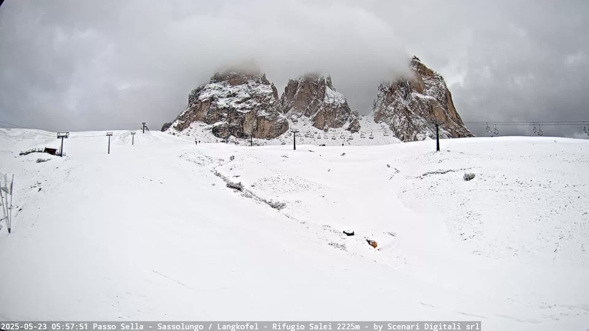 A snow-covered mountain slope with ski lifts and three rocky peaks partially obscured by clouds under an overcast sky.