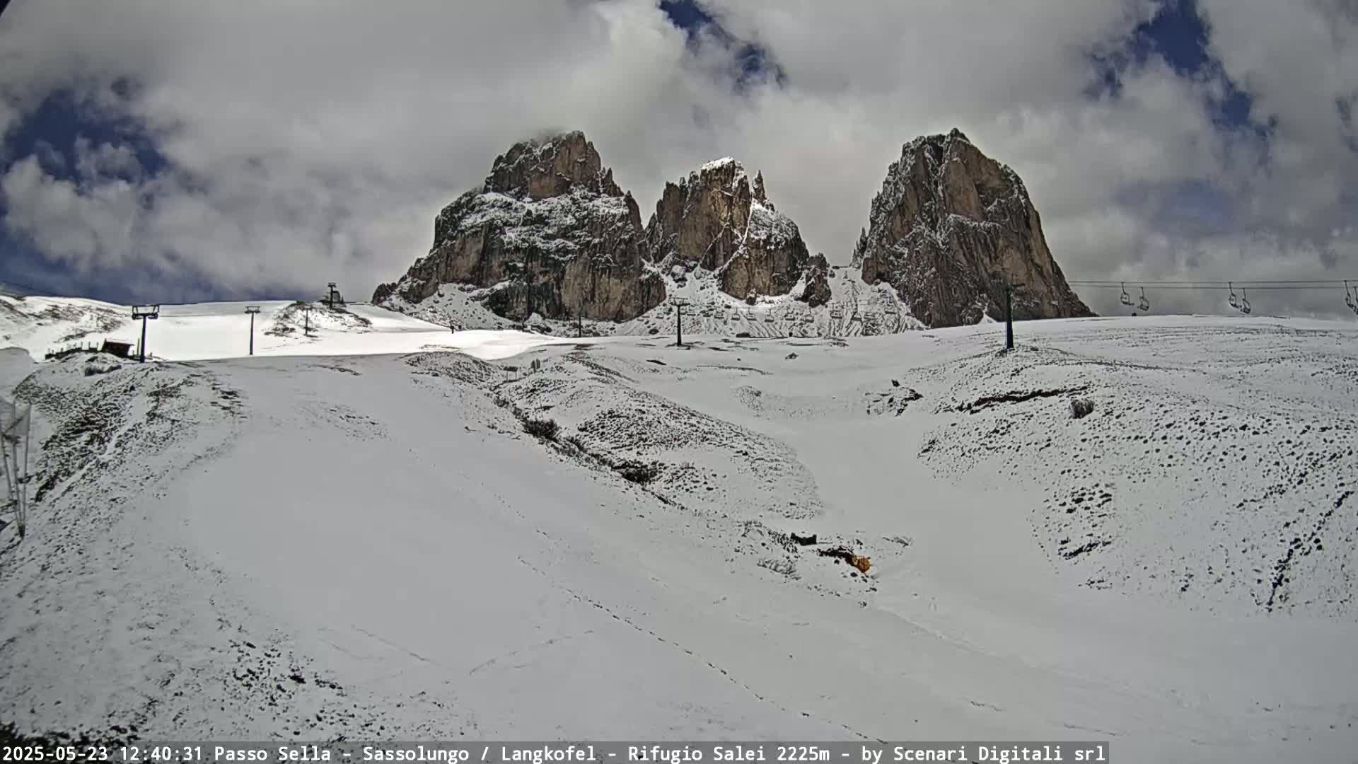 A snow-covered mountain landscape with three prominent rock formations under a cloudy sky, featuring ski lifts in the background.