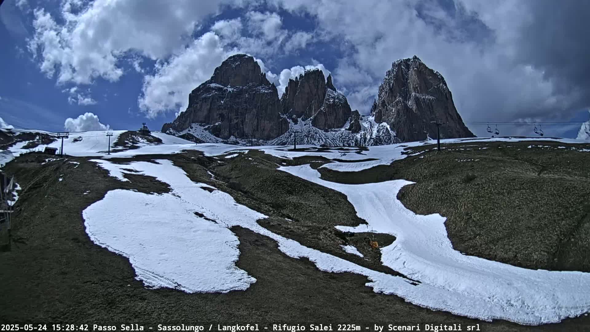 A partly cloudy day shows patches of snow on a mountainside with a ski lift and rocky, jagged peaks in the background.