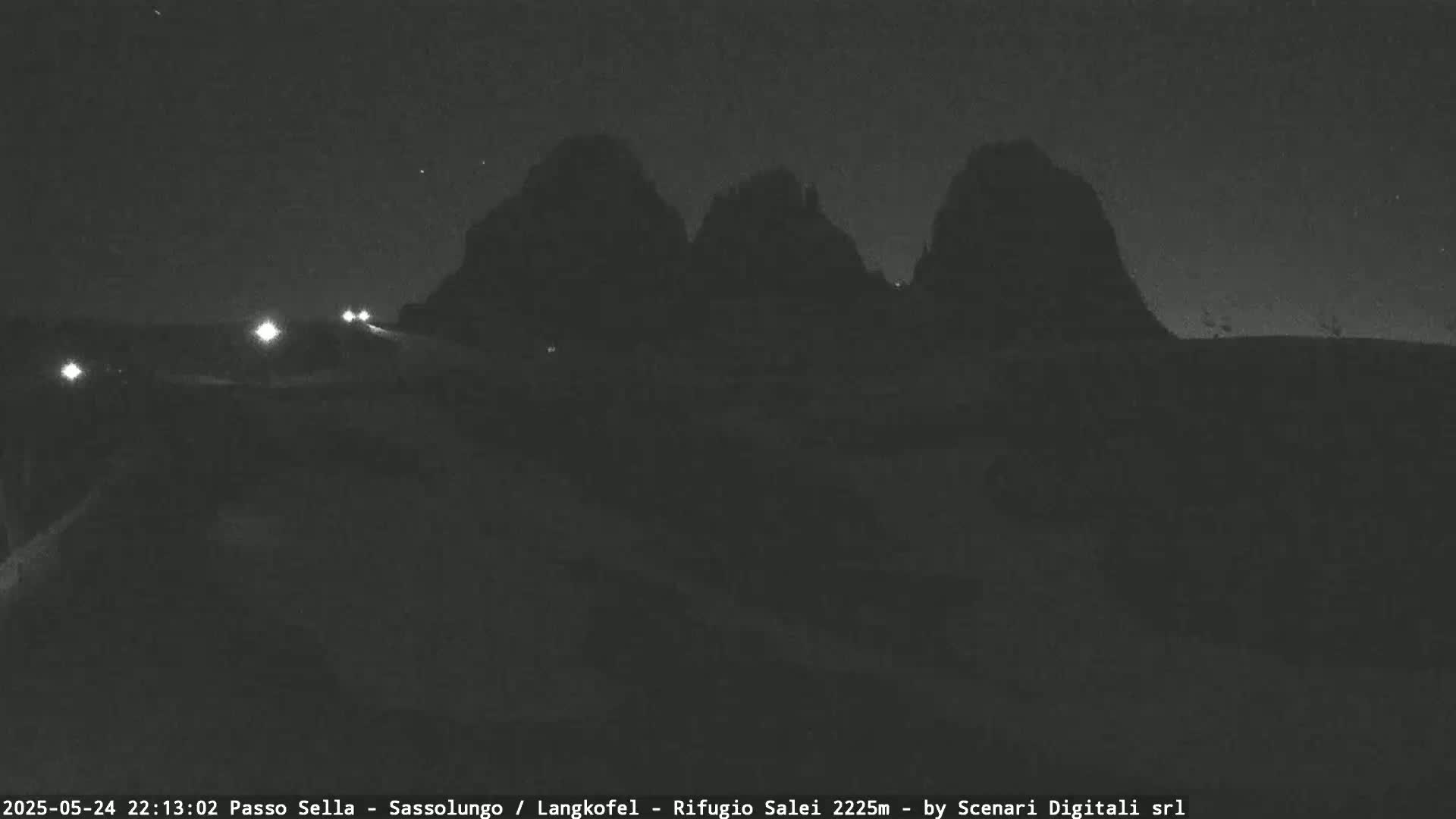 A nighttime view of a road with a few vehicle headlights visible, leading towards silhouetted mountains under a clear, starry sky.