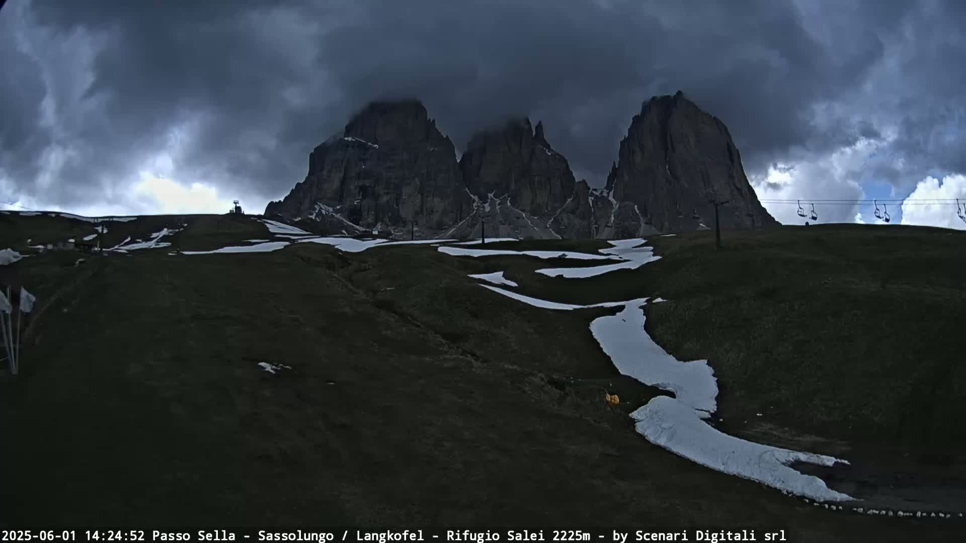 Dark, ominous clouds loom over snow-dusted, grassy hills and towering, jagged mountain peaks under a mostly cloudy sky.