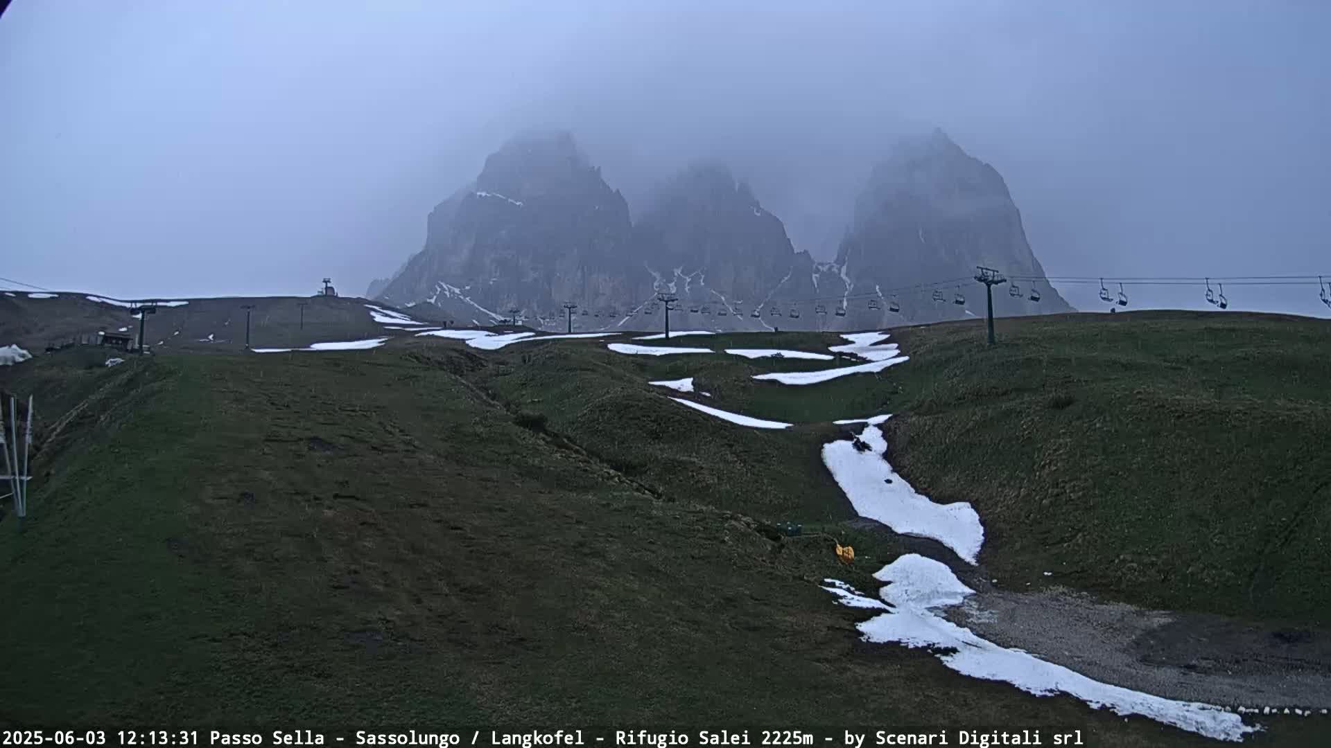 A chairlift runs across a grassy hillside dotted with patches of snow, in front of a misty mountain range.
