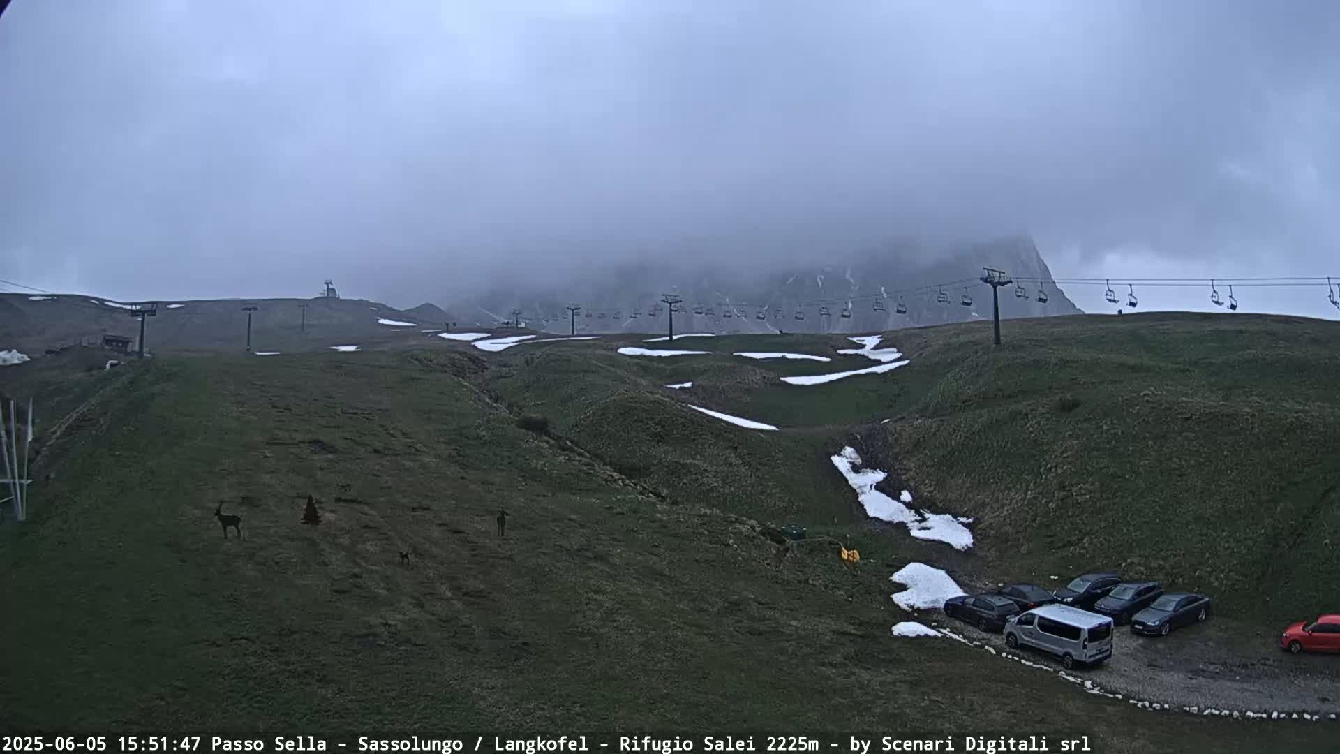 A foggy mountain landscape with a chairlift, patches of snow, and several cars parked near the base.