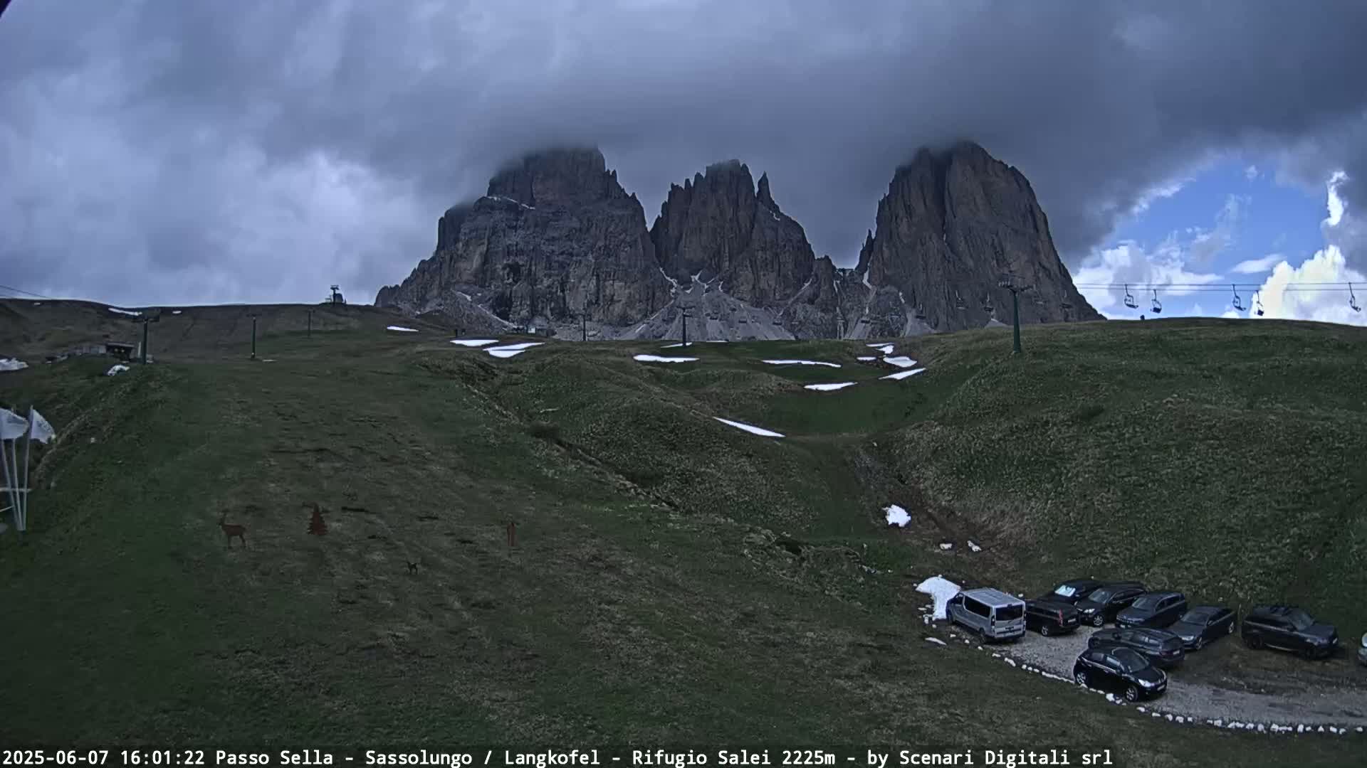 A mountain range is partially obscured by clouds over a grassy hillside with patches of snow and a small parking lot of vehicles.