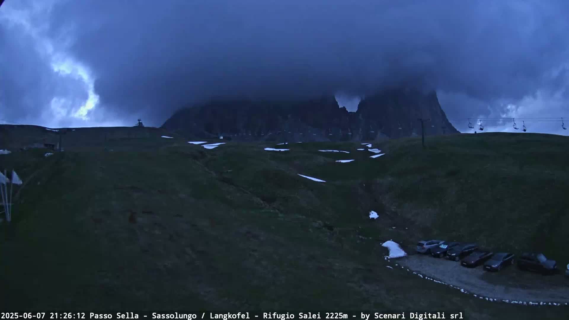 A mountain range is shrouded in dark, stormy clouds, with a few patches of snow visible on the ground below, and several cars parked in a lot in the foreground.