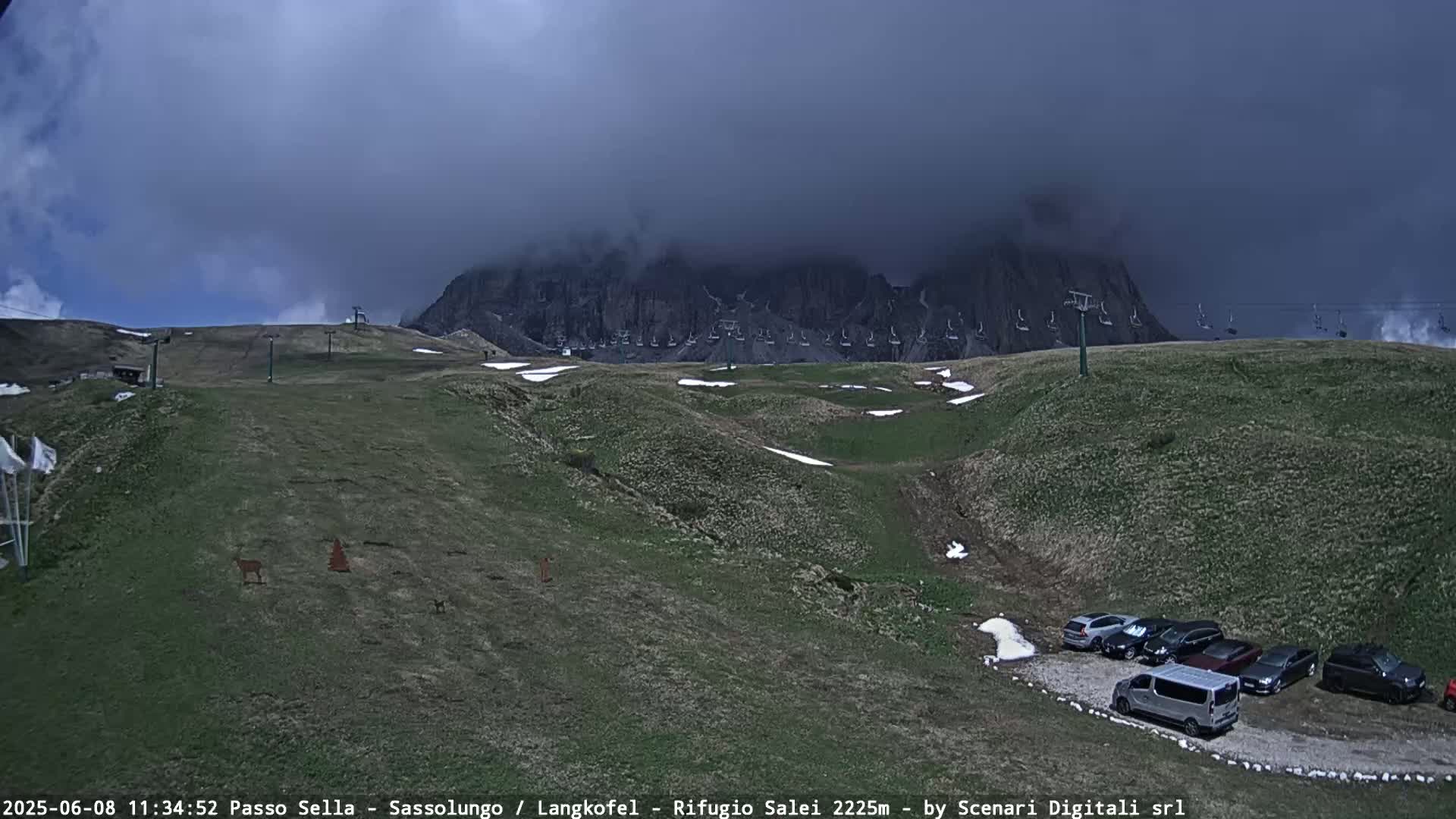 A mountain range partially obscured by dark clouds is visible above a grassy hillside with a parking lot and chairlifts, and small patches of snow remain on the ground.