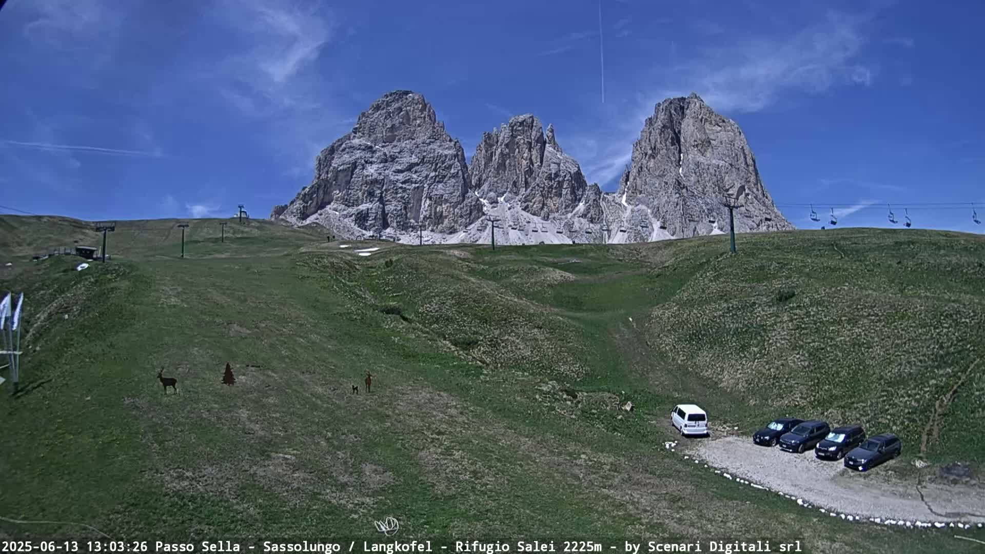 A grassy mountainside with a chairlift and several cars parked near a rocky mountain peak under a clear blue sky.