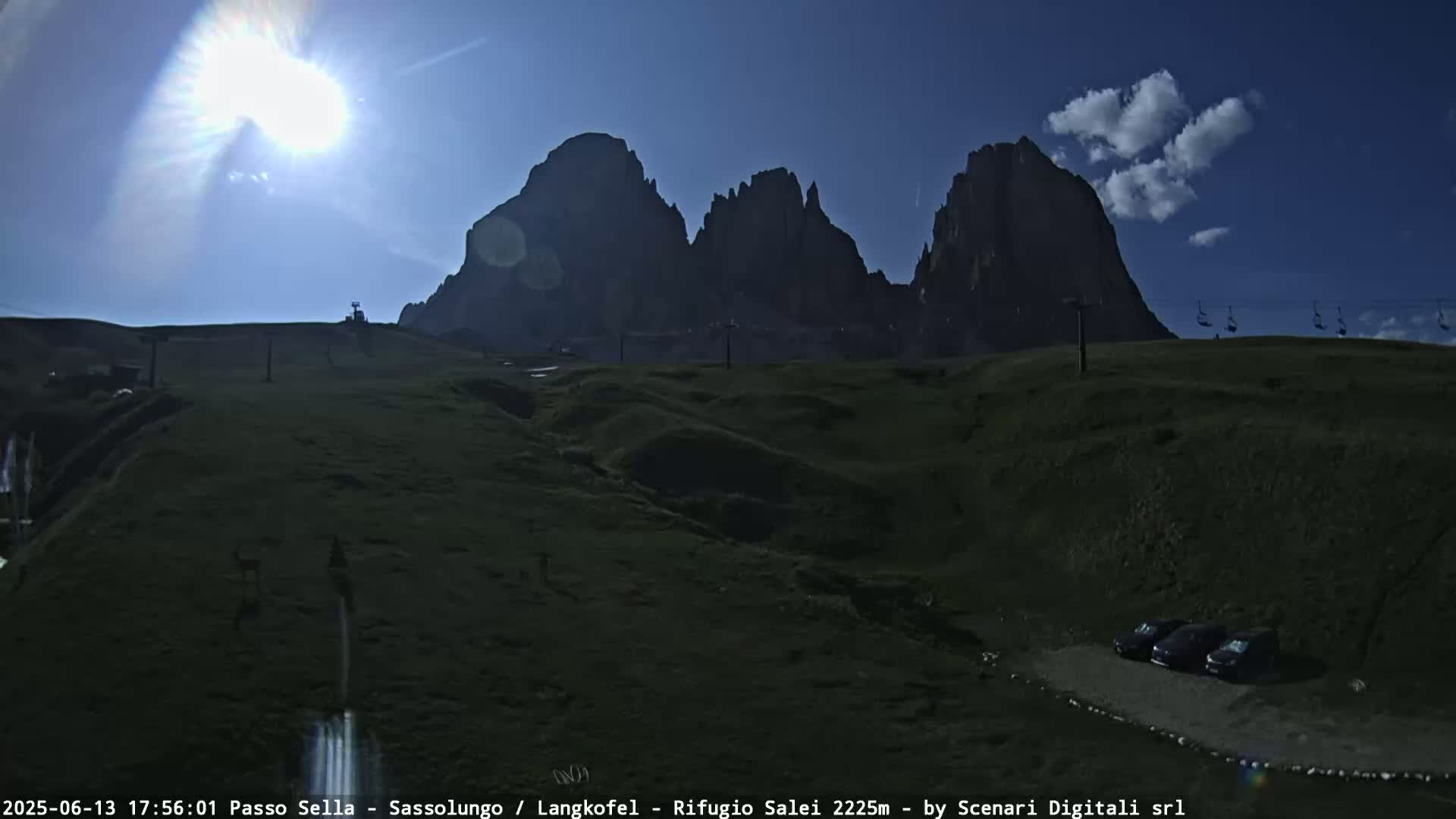 A sunny day reveals a grassy mountain landscape with a few cars parked near a chairlift, and a range of dark, jagged mountains in the background.
