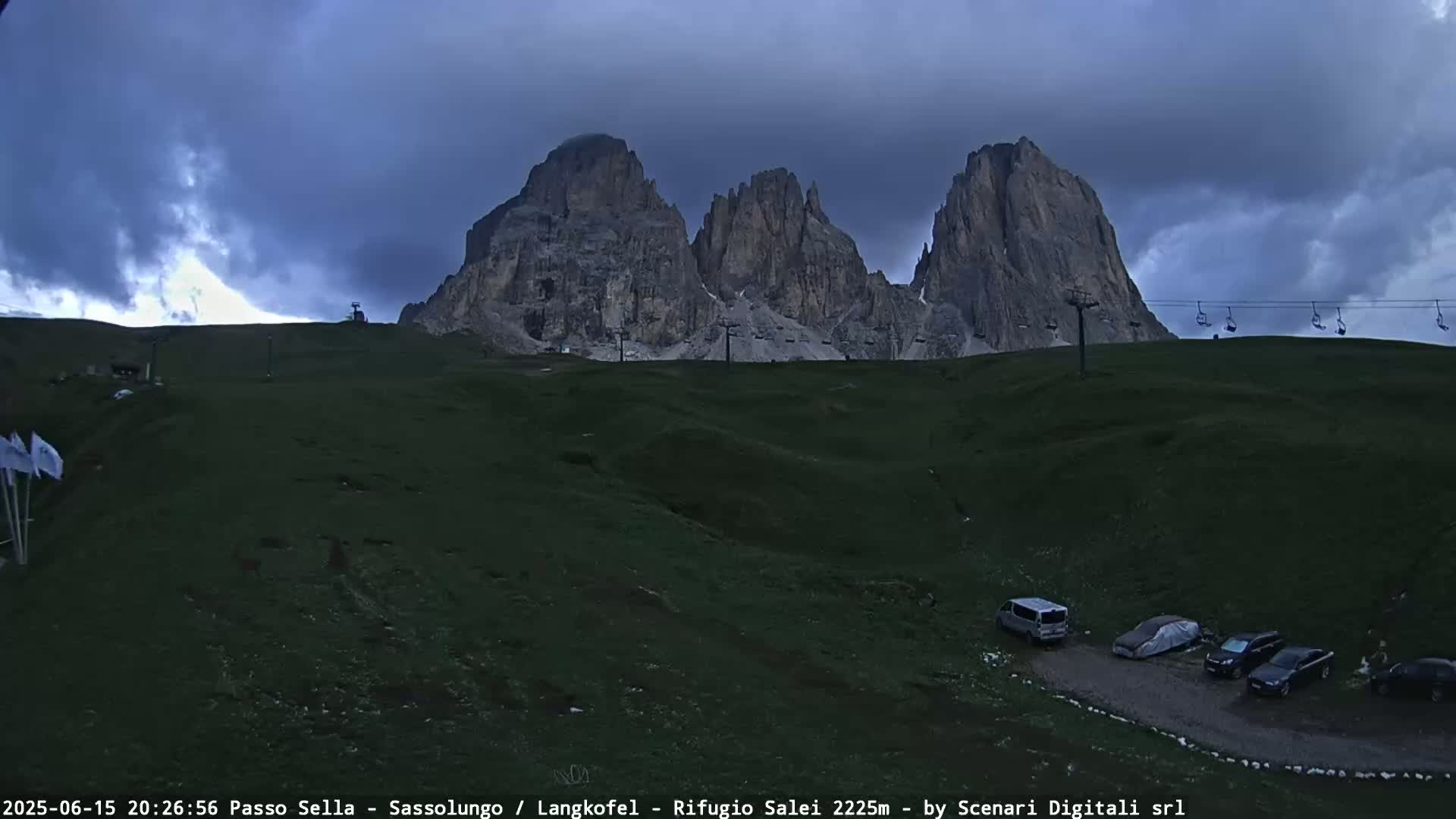 A grassy hillside with a few parked cars sits beneath a large, dark, rocky mountain range under a cloudy sky.