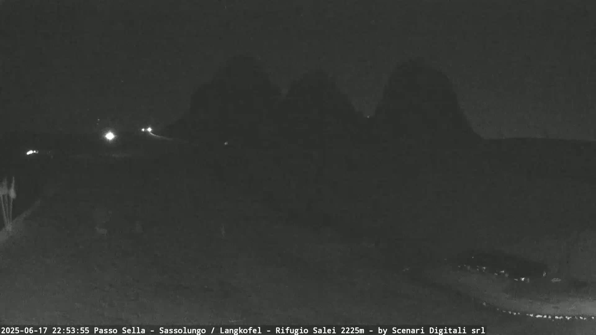A nighttime, low-light image shows dark, mountainous terrain under a clear, dark sky with a few distant vehicle headlights visible.