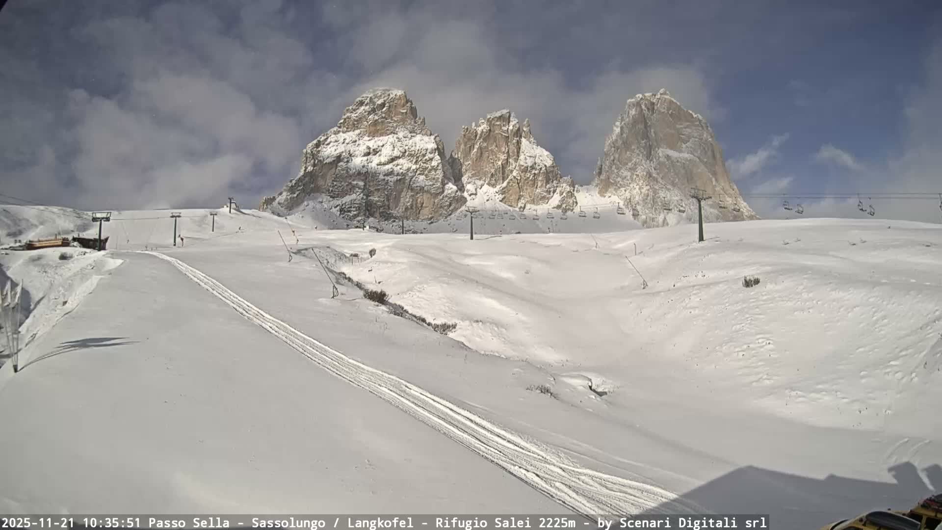 The image shows a vast snow-covered mountain ski resort on a partly cloudy winter day, featuring three prominent, rugged peaks in the background, extensive ski slopes with visible tracks, and multiple empty chairlifts spanning the terrain.