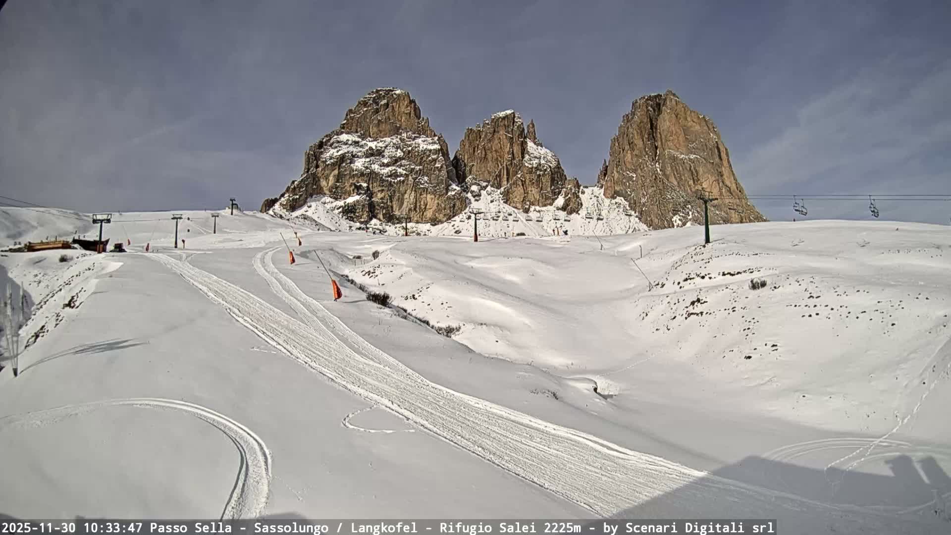 A snow-covered ski slope with ski tracks and multiple ski lifts is visible against a backdrop of rugged, snow-dusted mountains under a partly cloudy sky.