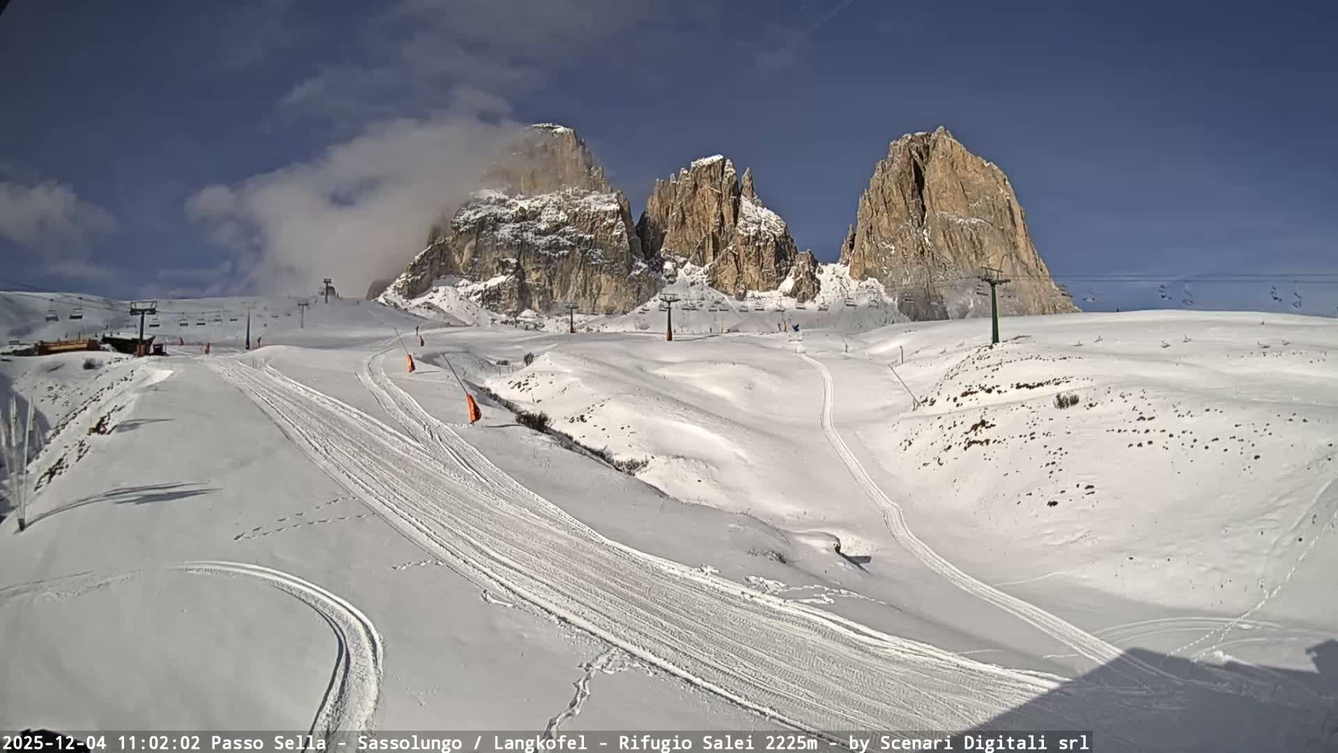 A vast, snow-covered ski resort with groomed slopes, chairlifts, and multiple ski tracks sprawls before majestic rocky mountain peaks partially shrouded in clouds, all under a clear blue sky with scattered white clouds.