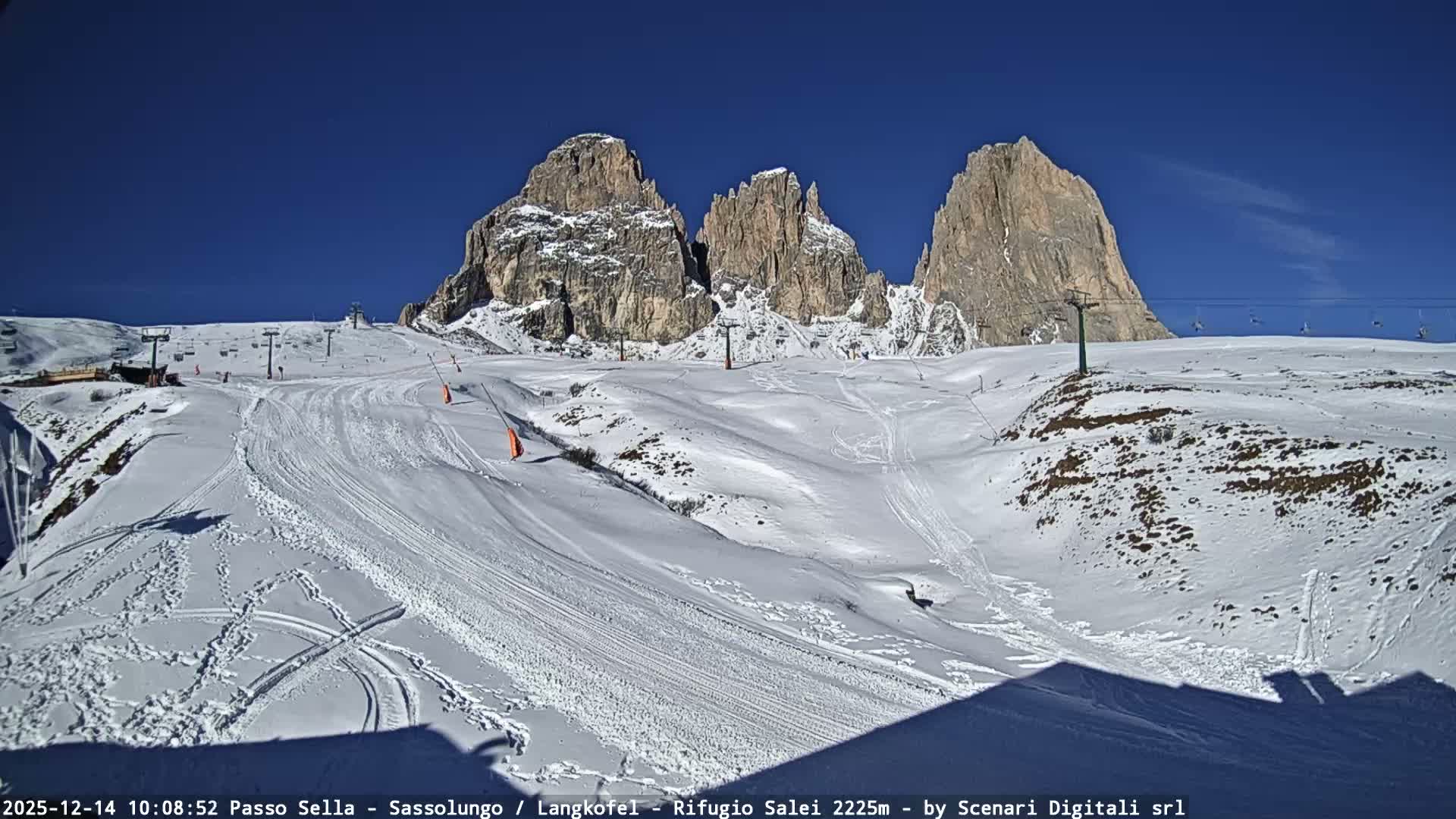 A vast, snow-covered ski resort with groomed slopes, chairlifts, and multiple ski tracks sprawls before majestic rocky mountain peaks partially shrouded in clouds, all under a clear blue sky with scattered white clouds.