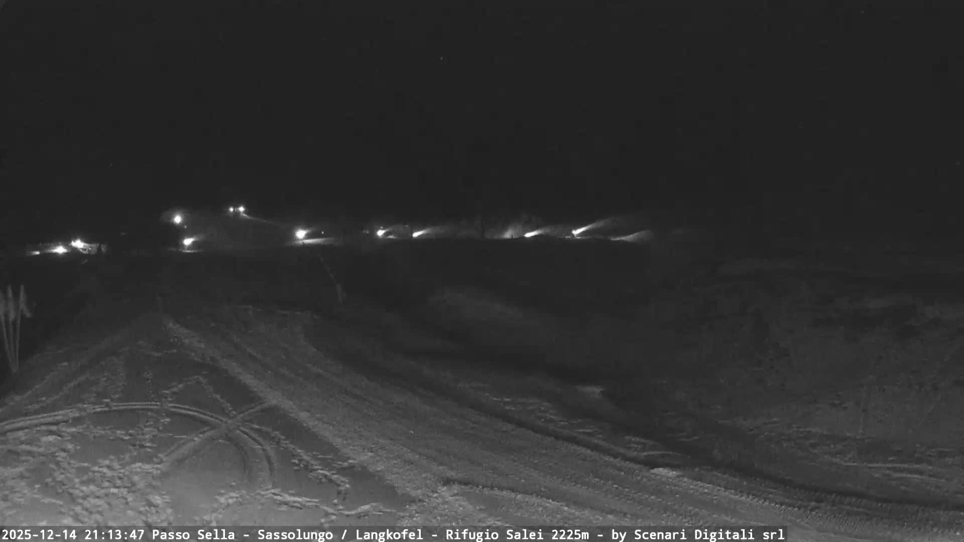 A vast, snow-covered ski resort with groomed slopes, chairlifts, and multiple ski tracks sprawls before majestic rocky mountain peaks partially shrouded in clouds, all under a clear blue sky with scattered white clouds.