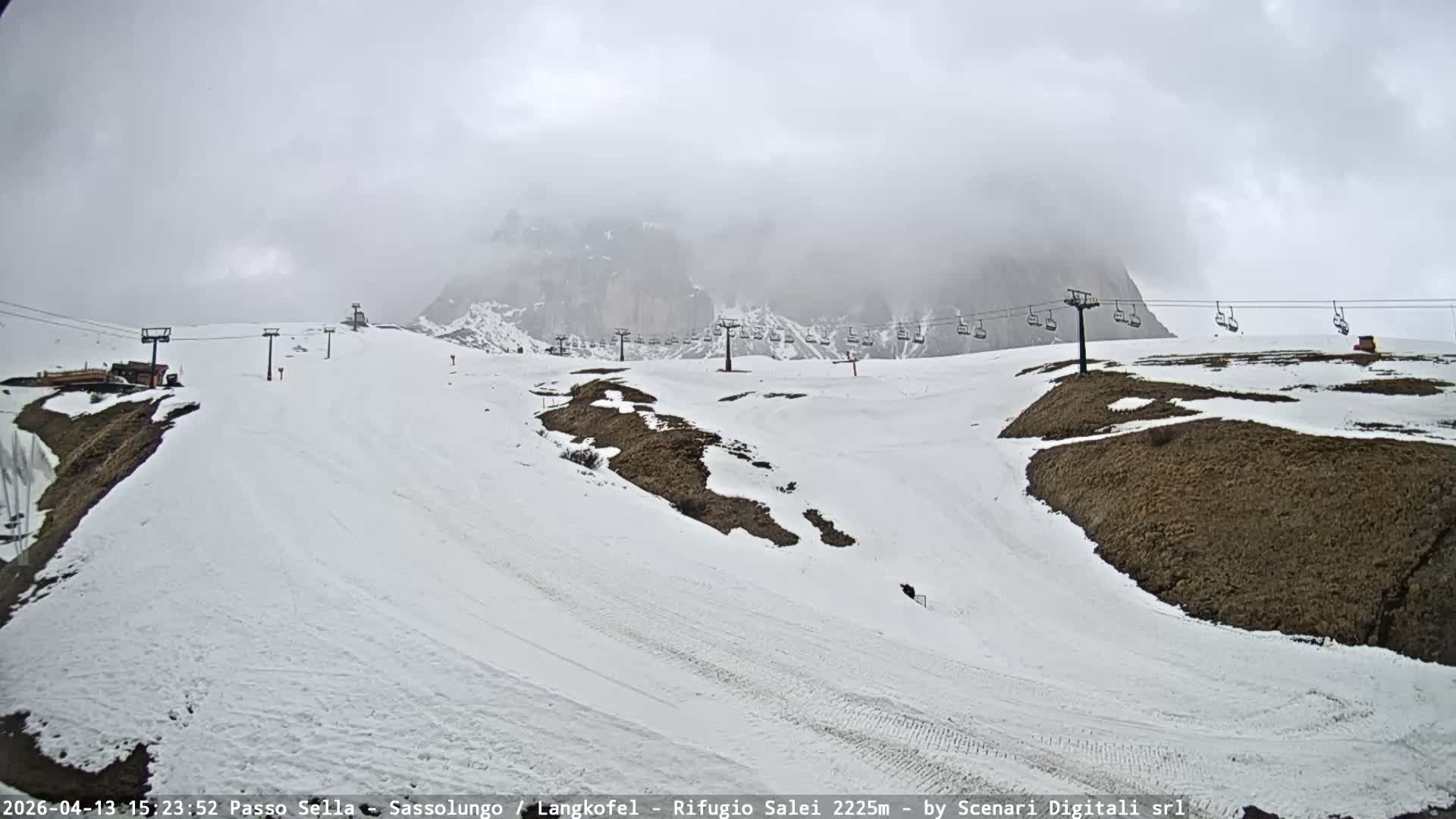 A vast, snow-covered ski resort with groomed slopes, chairlifts, and multiple ski tracks sprawls before majestic rocky mountain peaks partially shrouded in clouds, all under a clear blue sky with scattered white clouds.