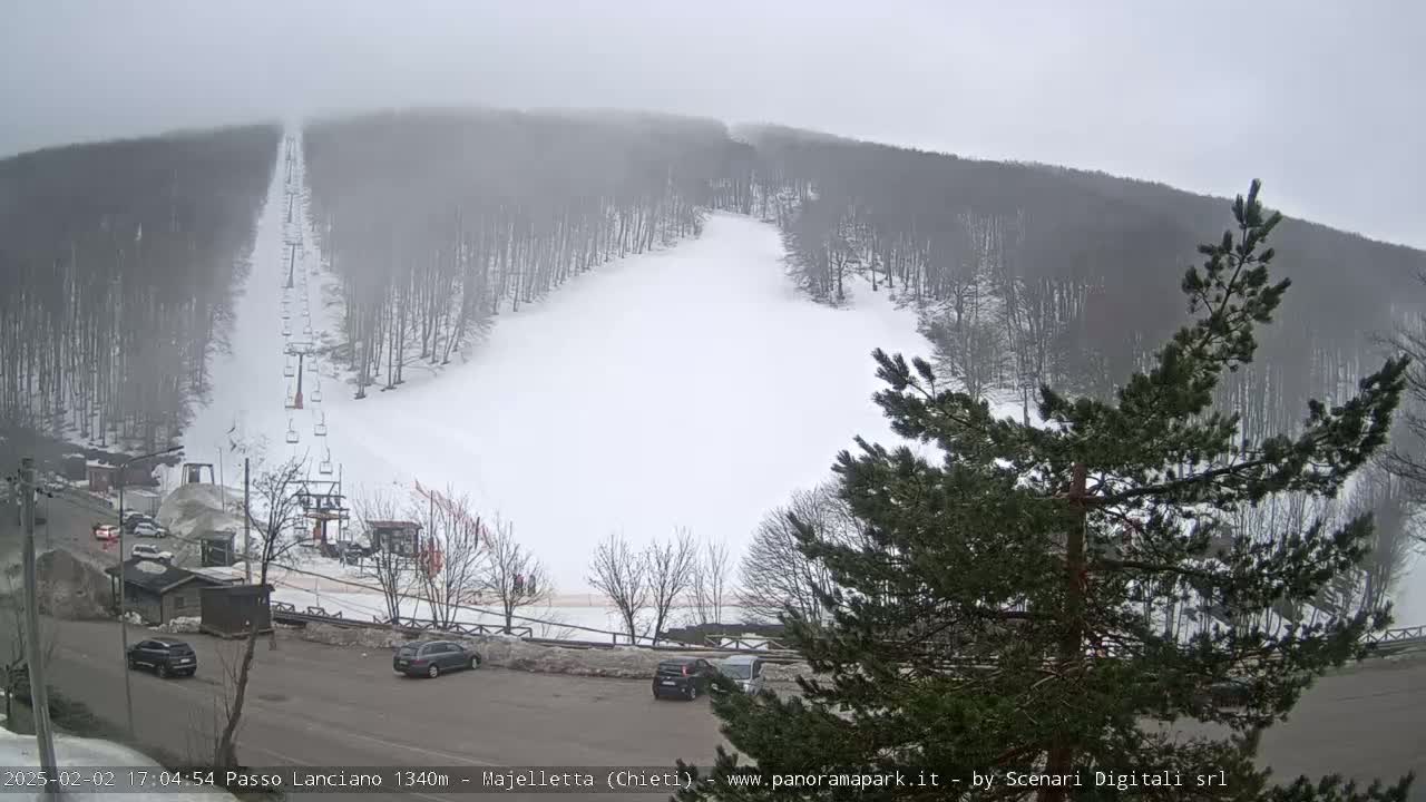 A snow-covered ski slope with a chairlift is visible through a foggy atmosphere, with several cars parked in a lot below.