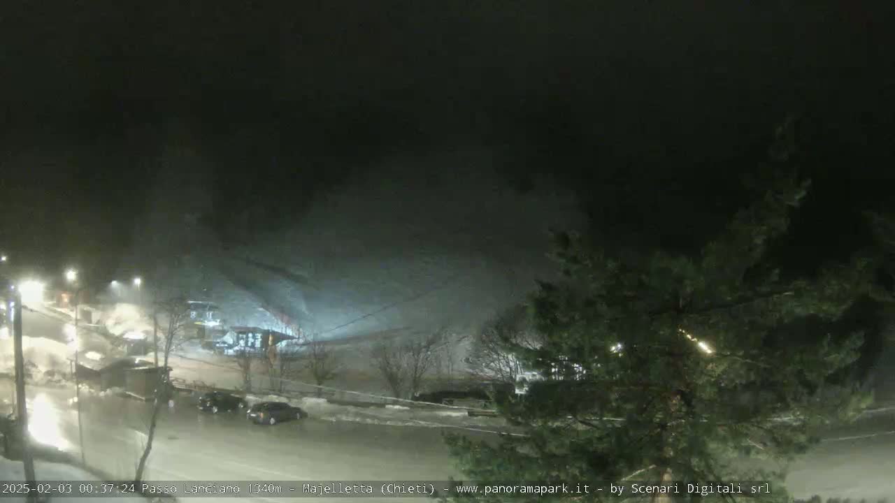 A nighttime, snowy scene shows a parking lot with a few cars, a road, and a snow-covered hill in the background under a dark, overcast sky.