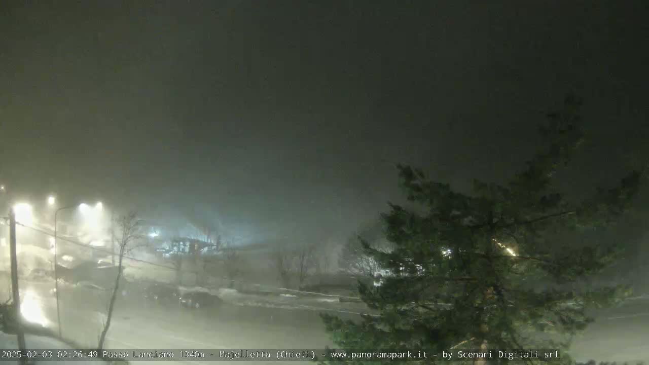 A nighttime scene of a snow-covered parking lot and road under heavy snowfall and fog.