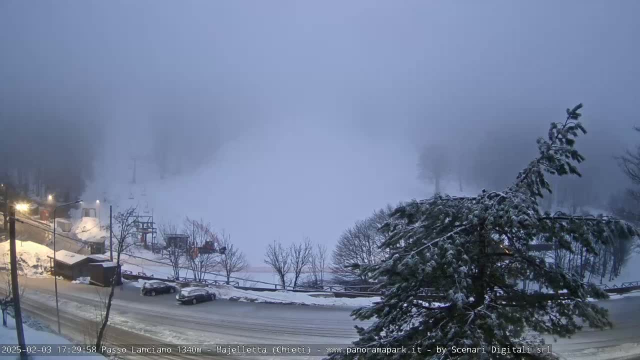 A snowy, foggy scene shows a parking lot with a few cars, some small buildings, and a ski lift in the background.