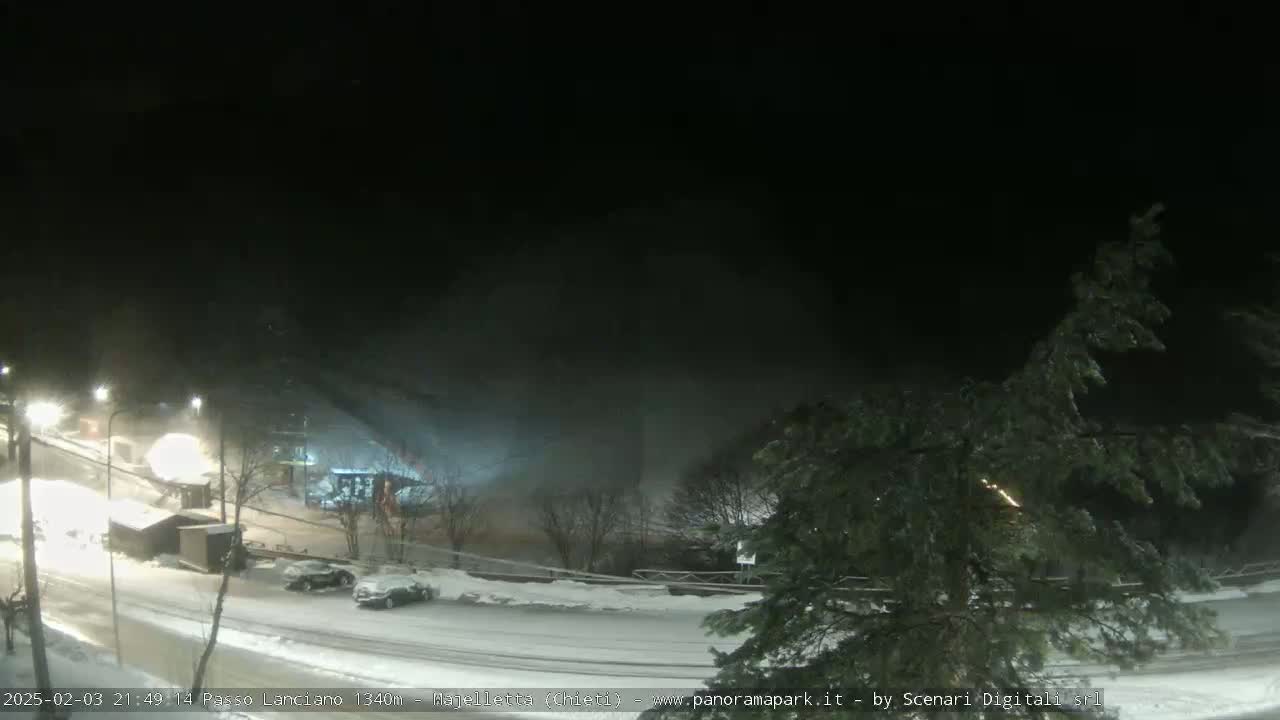 A snow-covered parking lot and road are visible at night, with some light snowfall occurring and a few vehicles parked.