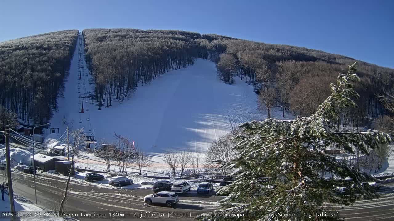 A snow-covered ski slope with a chairlift runs through a wooded hillside on a sunny day.